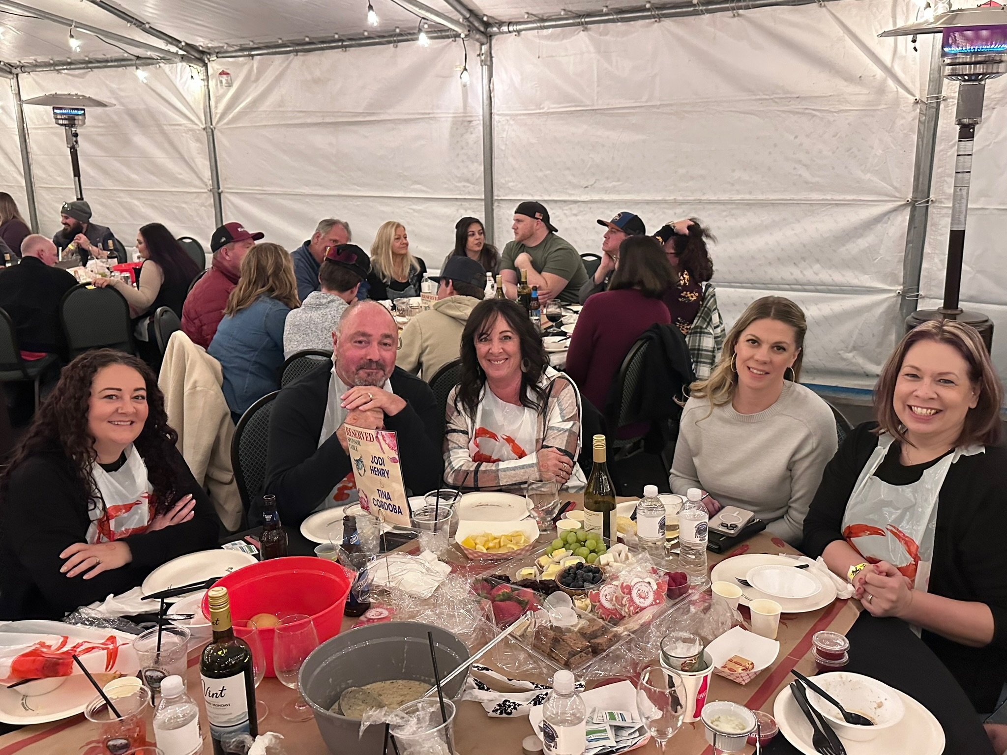People sitting at a large table inside a white tent engaging in a social gathering or celebration. The table has food, drinks, and various items on it, and the guests appear to be enjoying themselves.