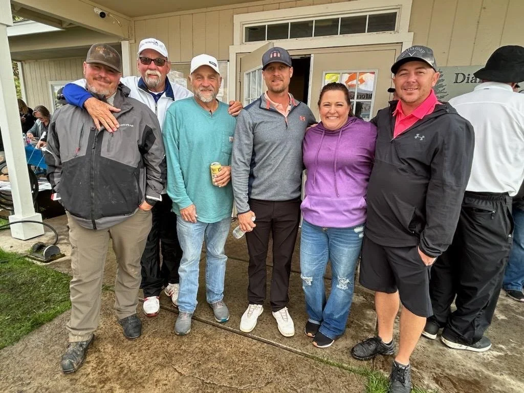 Group of six people standing together outdoors near a building, smiling for the camera. They are dressed in casual and sportswear, some holding drinks.