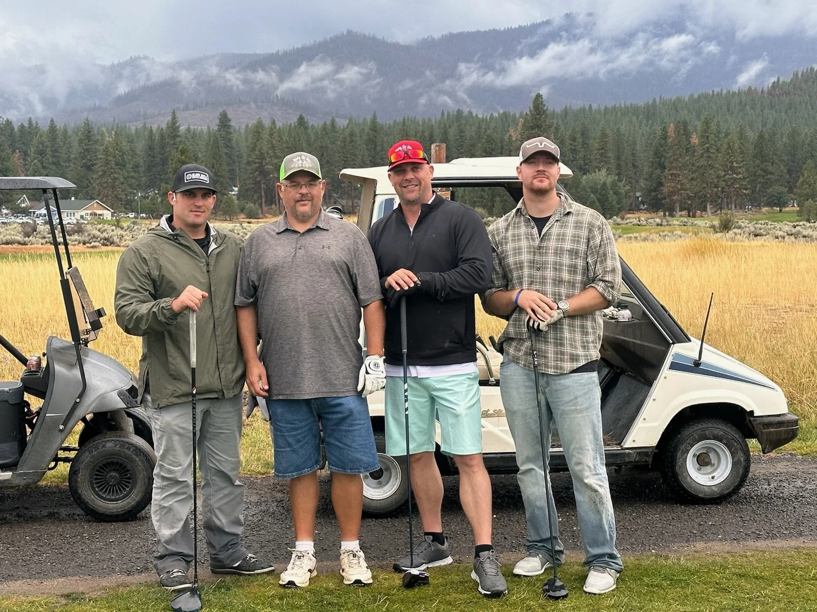 Four men standing on a golf course with two golf carts and a grassy field, mountains, and trees in the background.