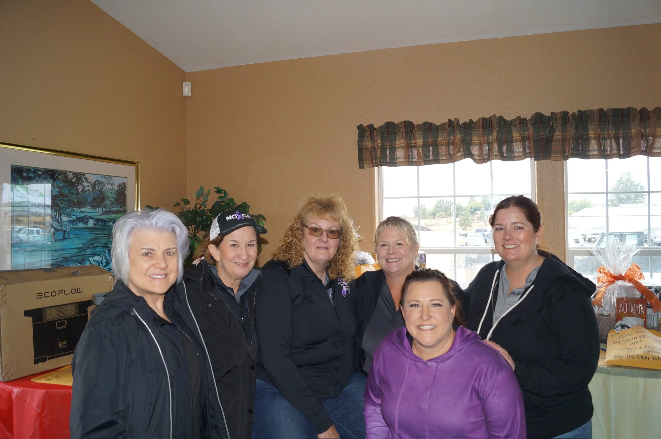 Group of women posing indoors at a gathering, smiling at the camera, with a window showing a parking lot outside.