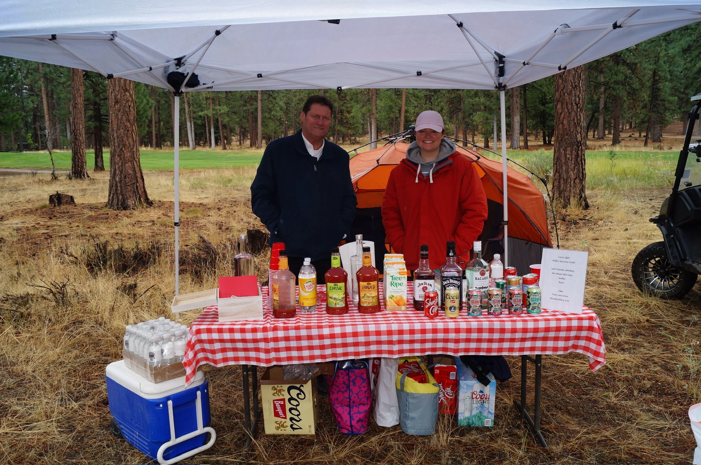 Two people standing behind a table with drinks and snacks at an outdoor event in a wooded area, under a white canopy tent.