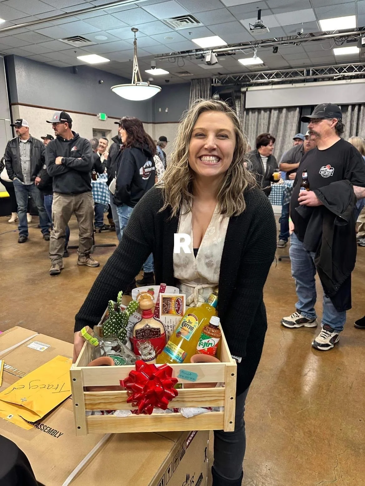 A woman with curly hair smiling at camera, standing behind a gift basket filled with various bottles and items, in a crowded indoor event space with people mingling in the background.