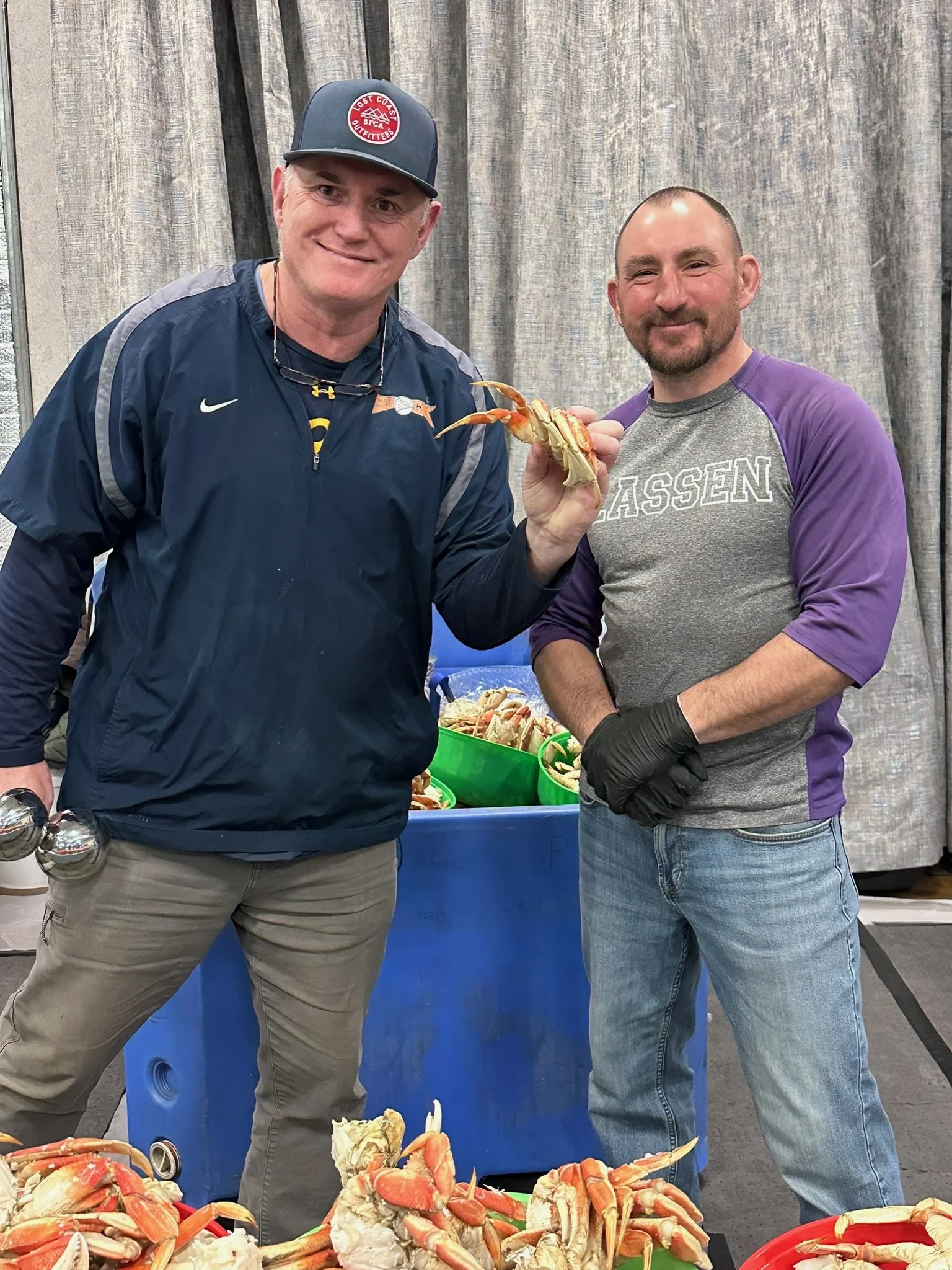 Two men standing next to a table full of cooked crab, holding a crab between them, indoors with gray curtains in the background. One man is wearing a blue jacket and a baseball cap, the other is wearing a gray and purple long sleeve shirt.