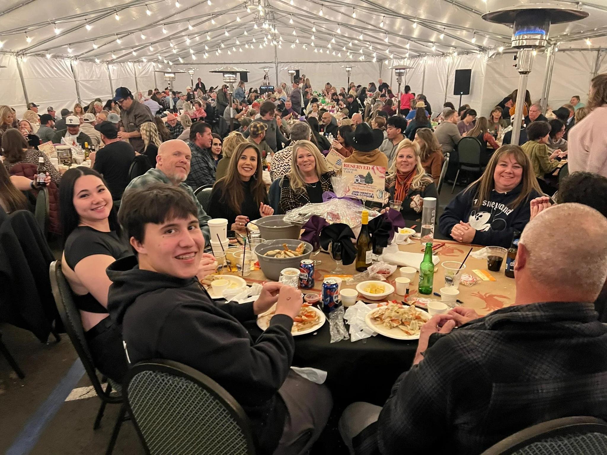 A large group of people gathered inside a spacious tent for a meal or celebration, with tables filled with food and drinks, and string lights hanging from the ceiling.