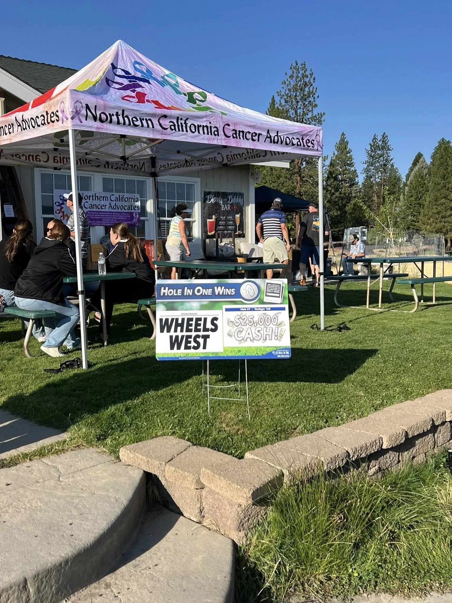 A tent with a purple and white roof and the words "Northern California Cancer Advocates" is set up outdoors during the day with people sitting and standing around it. A sign in front of the tent advertises a hole-in-one golf contest with a $25,000 cash prize, sponsored by Wheels West. There are trees and a building in the background.