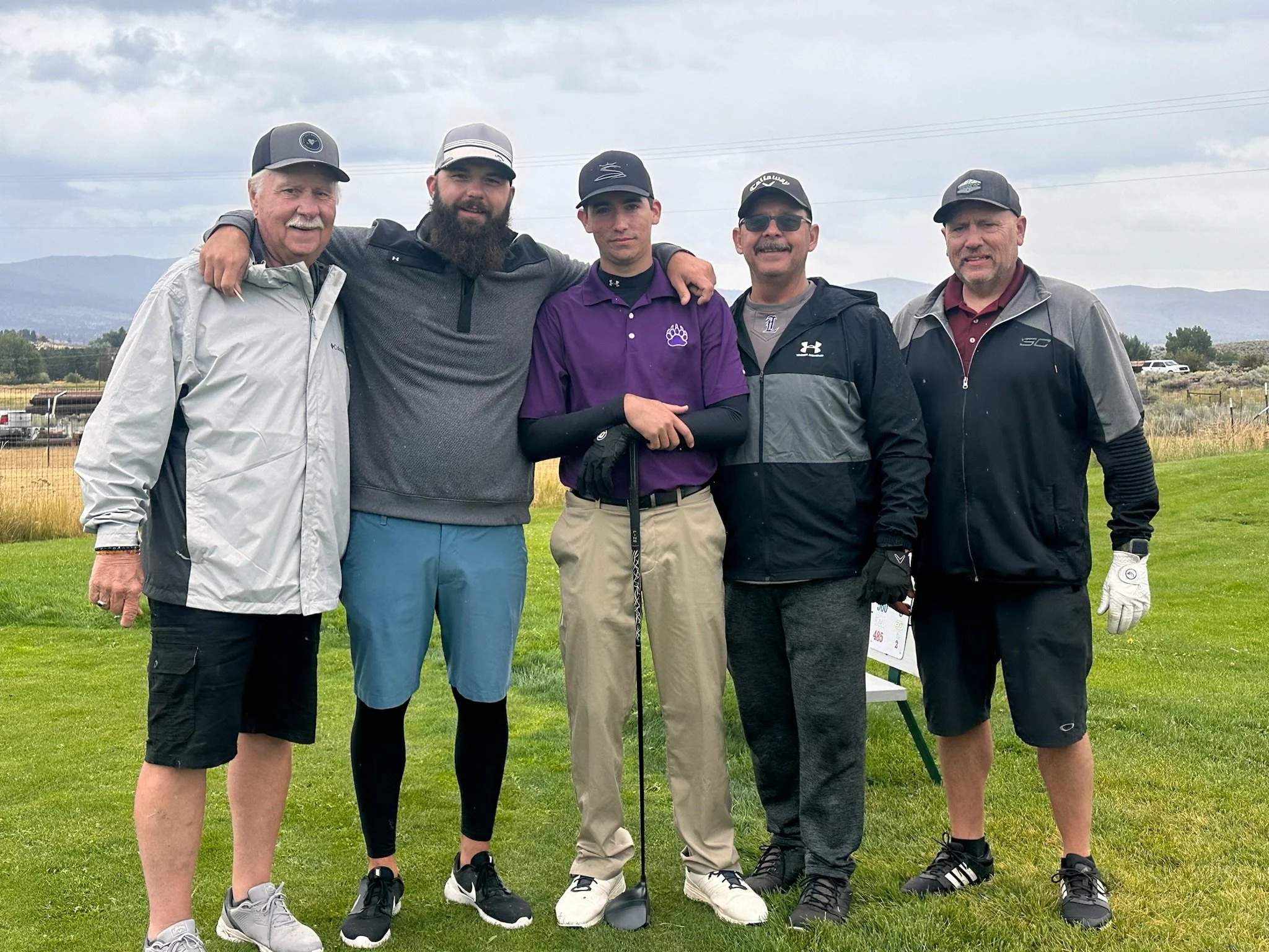 A group of six men standing outdoors on a golf course, with mountains and cloudy sky in the background, dressed in golf attire.