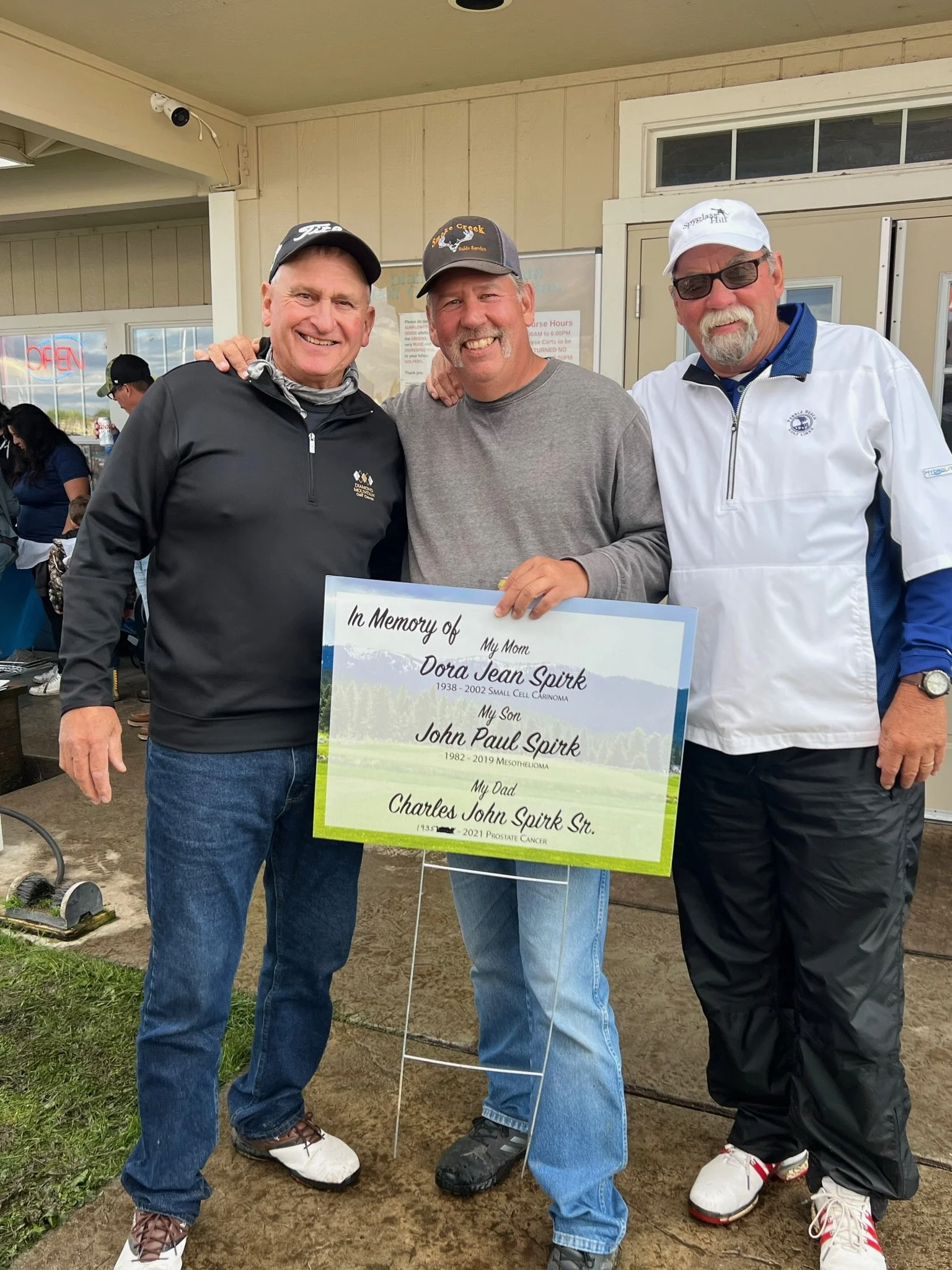 Three men standing outdoors in front of a building, smiling, with a large memorial sign in front of them. The sign reads: 'In Memory of My Mom Dora Jean Spirk 1938-2002 Small Cell Carinoma,' 'My Son John Paul Spirk 1982-2019 Mesothelioma,' 'My Dad Ch