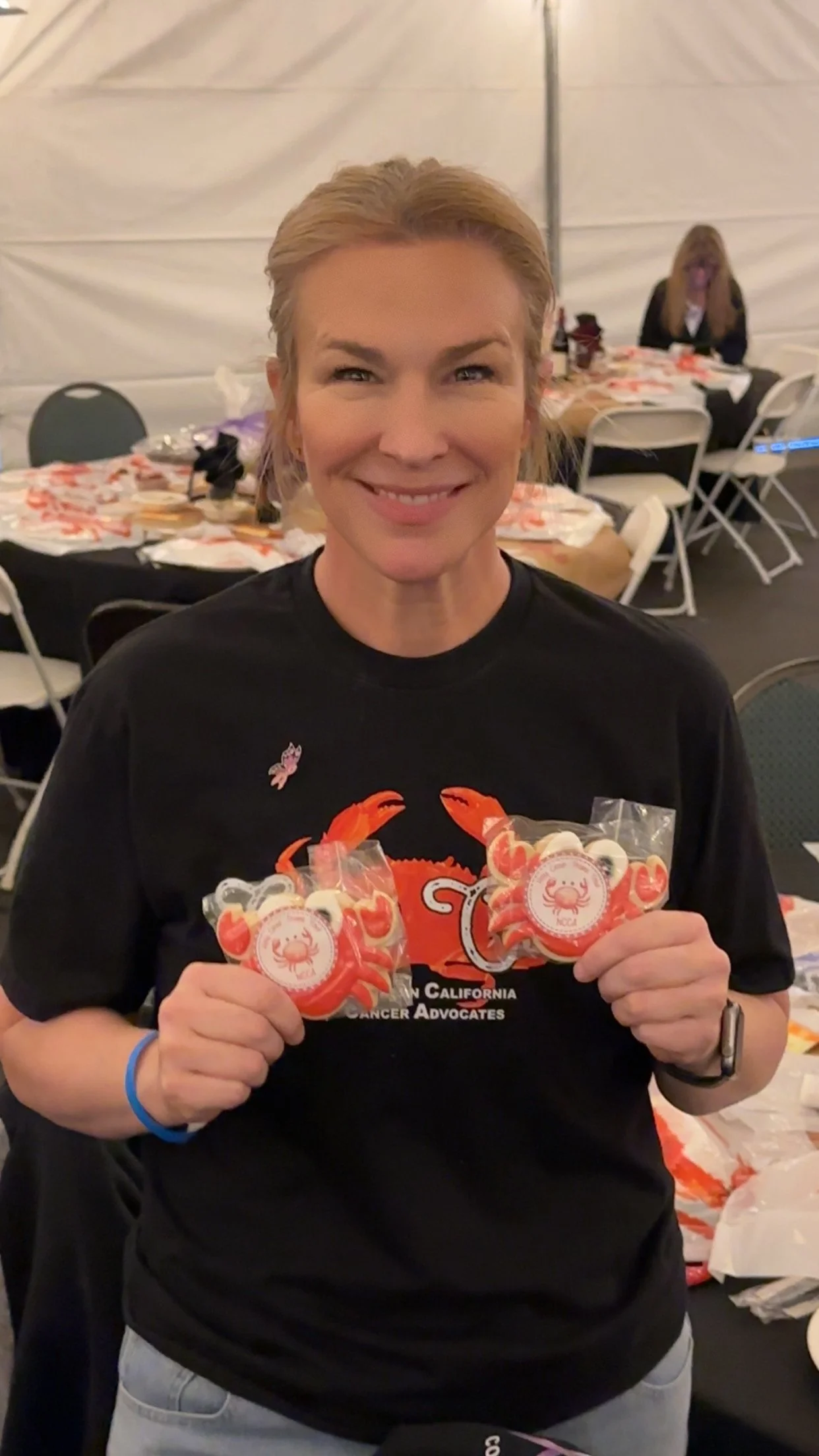 A woman smiling and holding small plastic bags with crab illustrations on them at a gathering, with round tables, chairs, and another woman in the background.