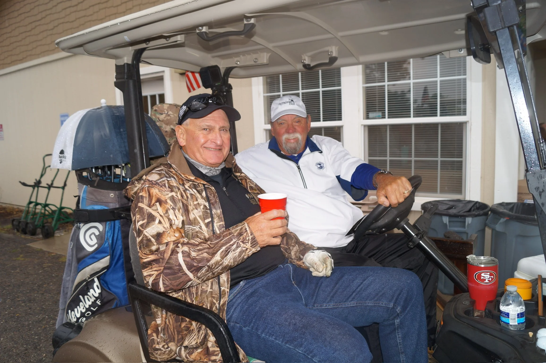 Two older men sitting in a golf cart, smiling, with one holding a red cup, outside a building with large windows and a few utility items nearby.