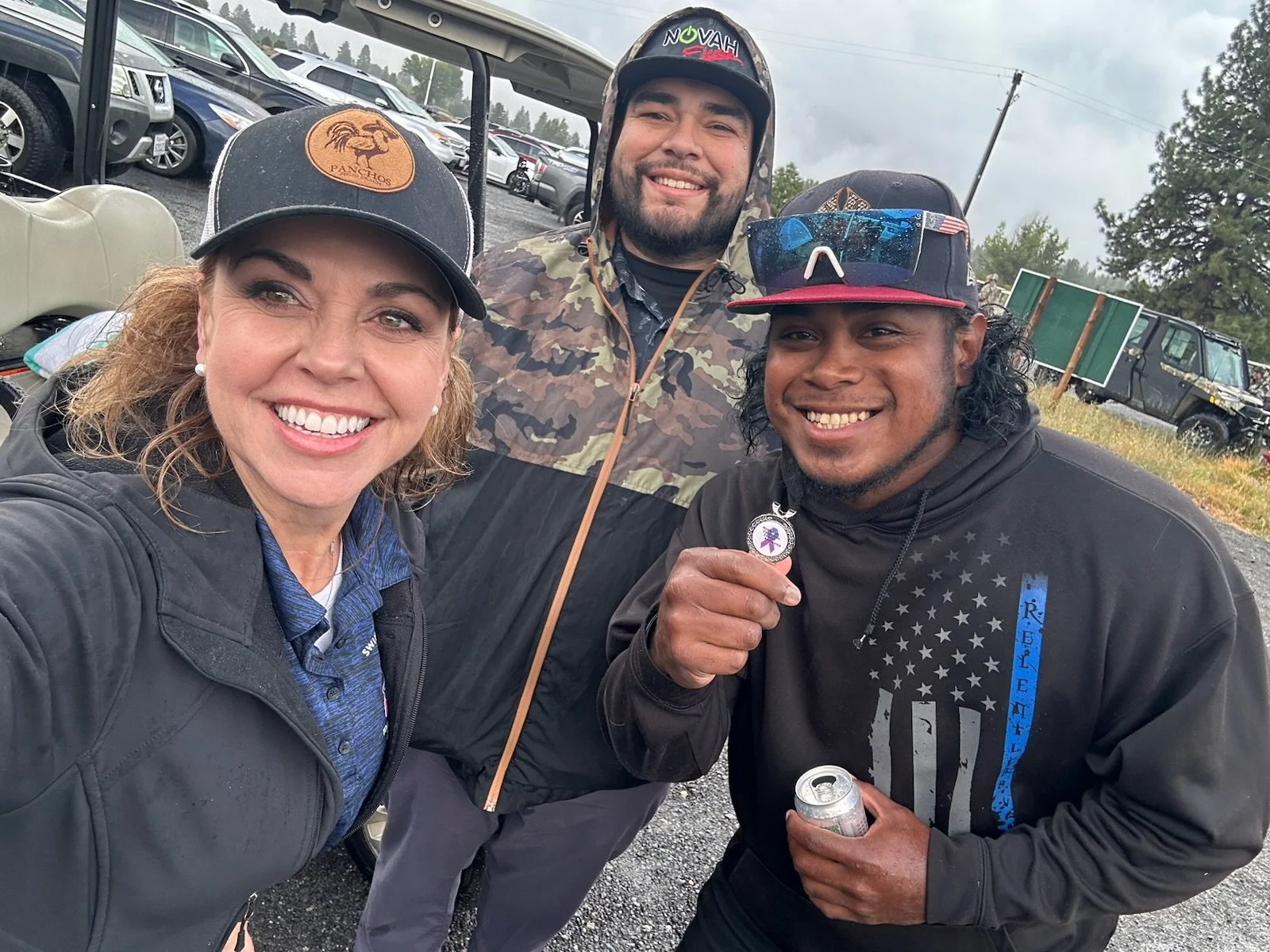 A group of three people smiling and taking a selfie outdoors in a parking lot on a cloudy day. They are dressed casually, and two are wearing hats and hoodies. Behind them are vehicles and some trees.