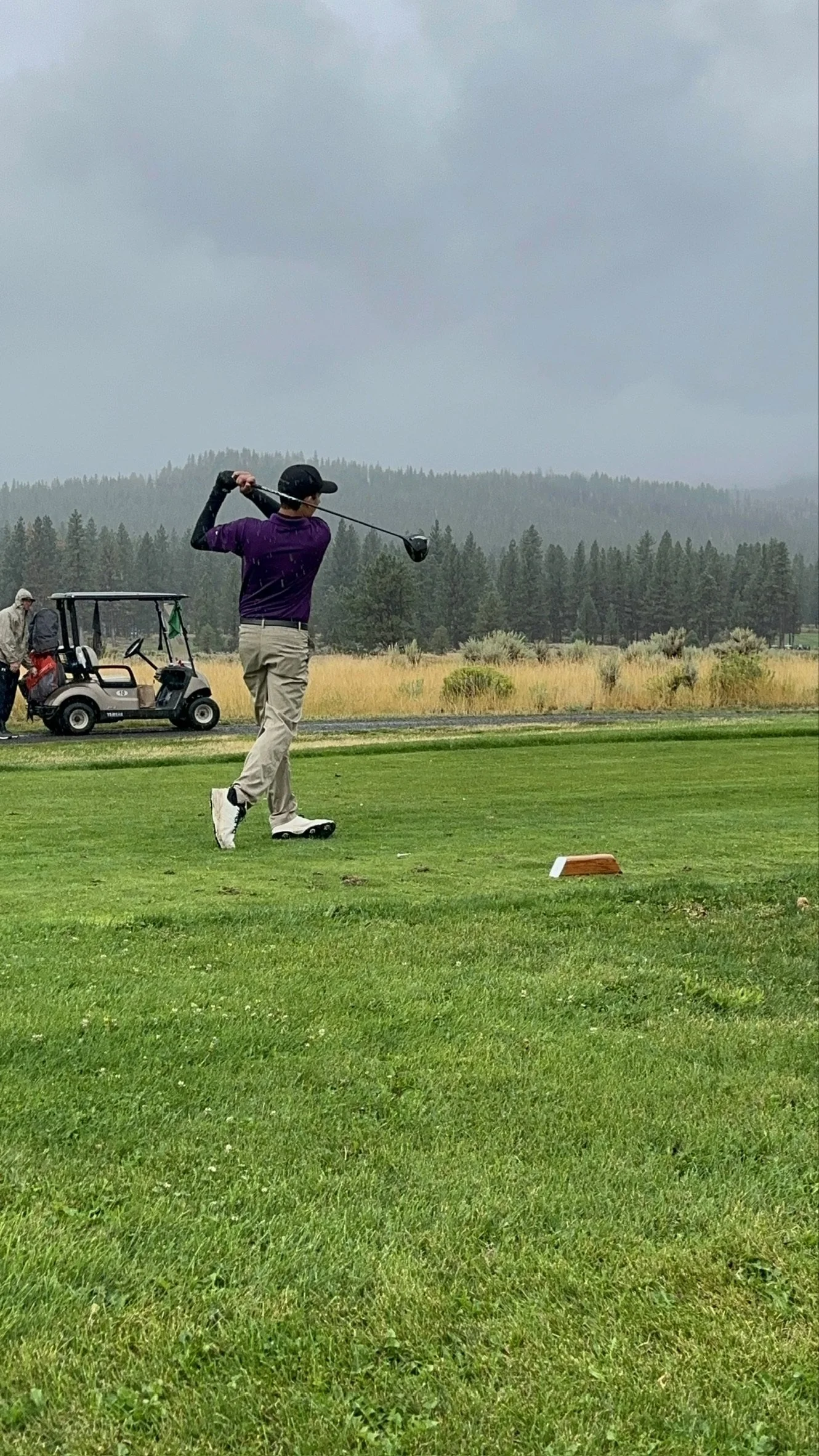 A person in a purple shirt and beige pants playing golf, swinging a golf club on a grassy golf course with trees and hills in the background, and another person near a golf cart.