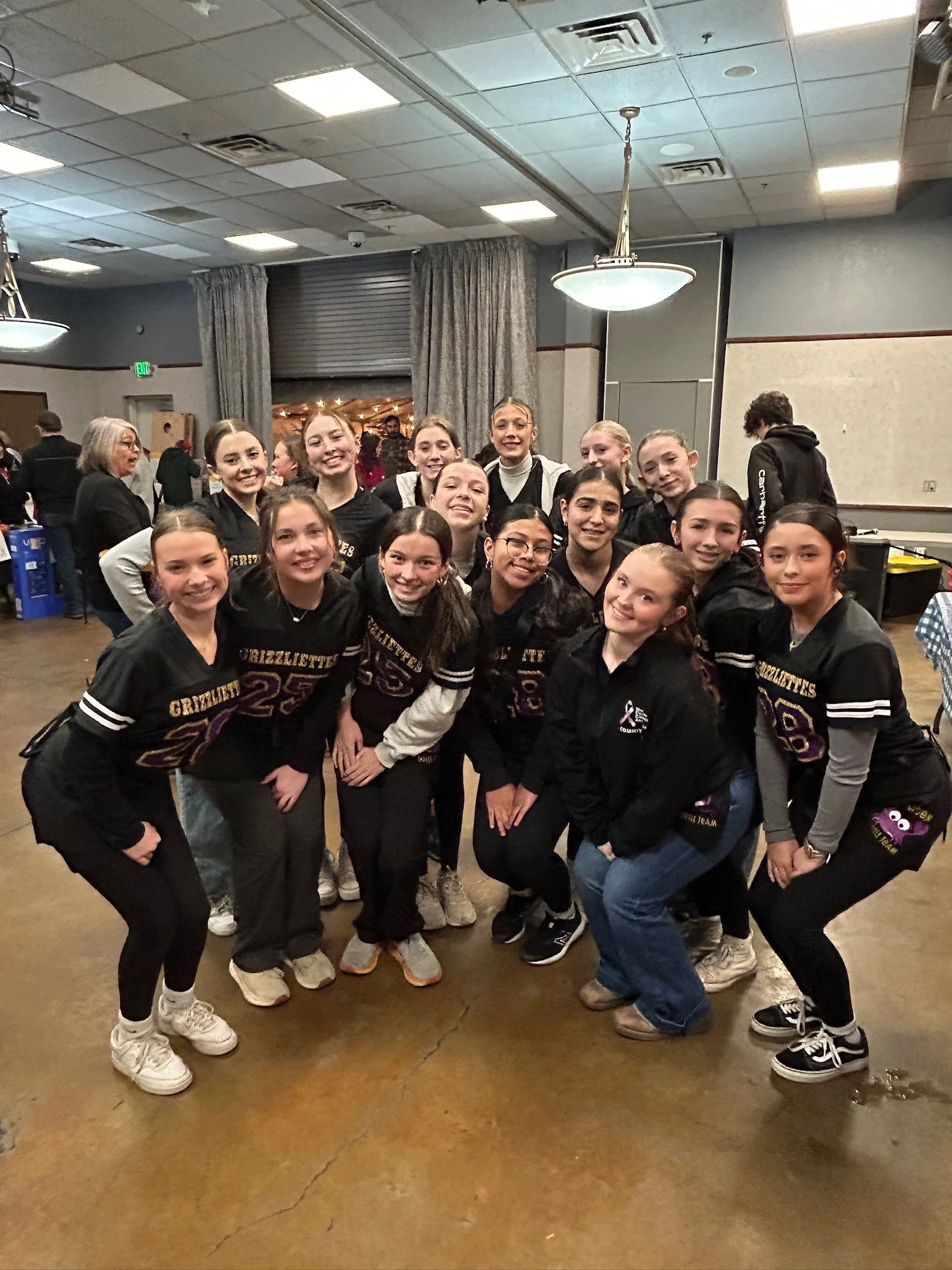 A group of young girls and women in matching black cheerleading uniforms posing indoors, smiling at the camera in front of a gray curtain and a closed window with metallic blinds.