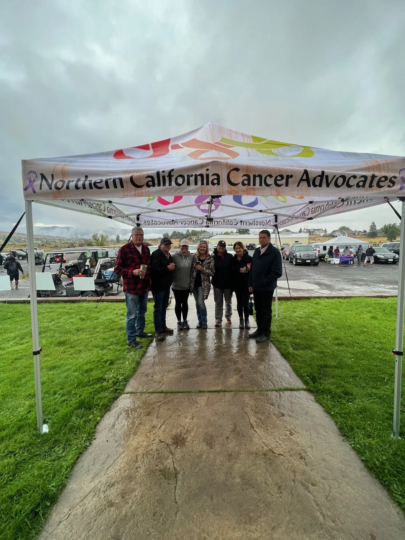 Group of six people standing in front of a Northern California Cancer Advocates tent at an outdoor event on a cloudy day, with parked cars and a grassy area in the background.