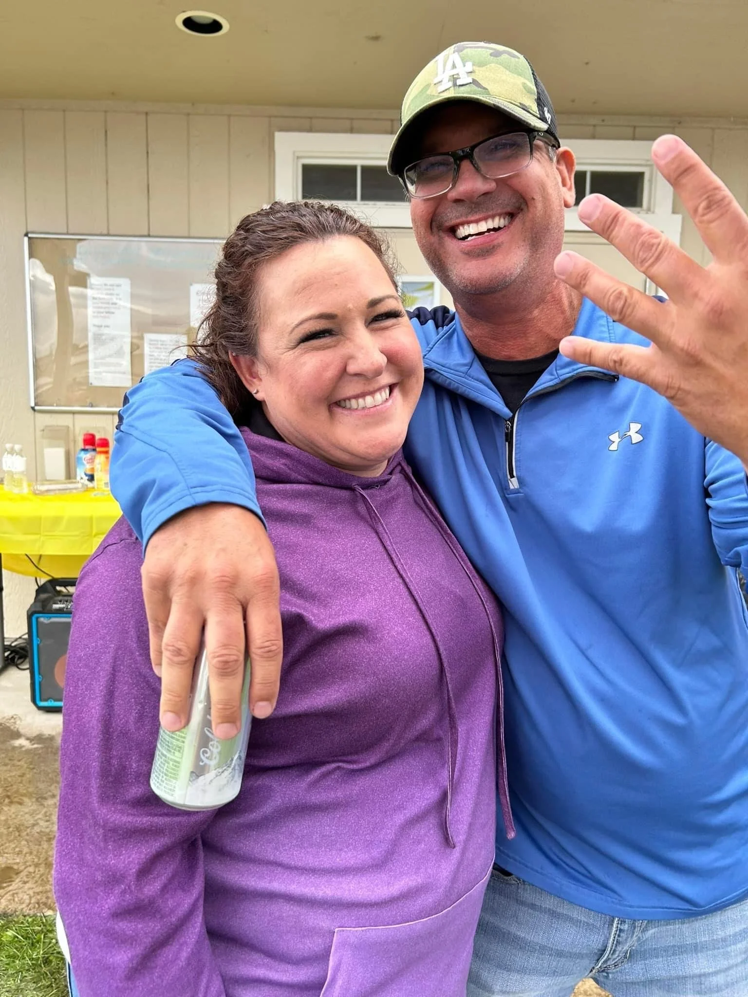 A smiling man wearing glasses, a camouflage cap, and a blue jacket is hugging a woman with a purple hoodie. The man is holding a water bottle in his hand. They are indoors with a bulletin board and various bottles on a yellow table in the background.