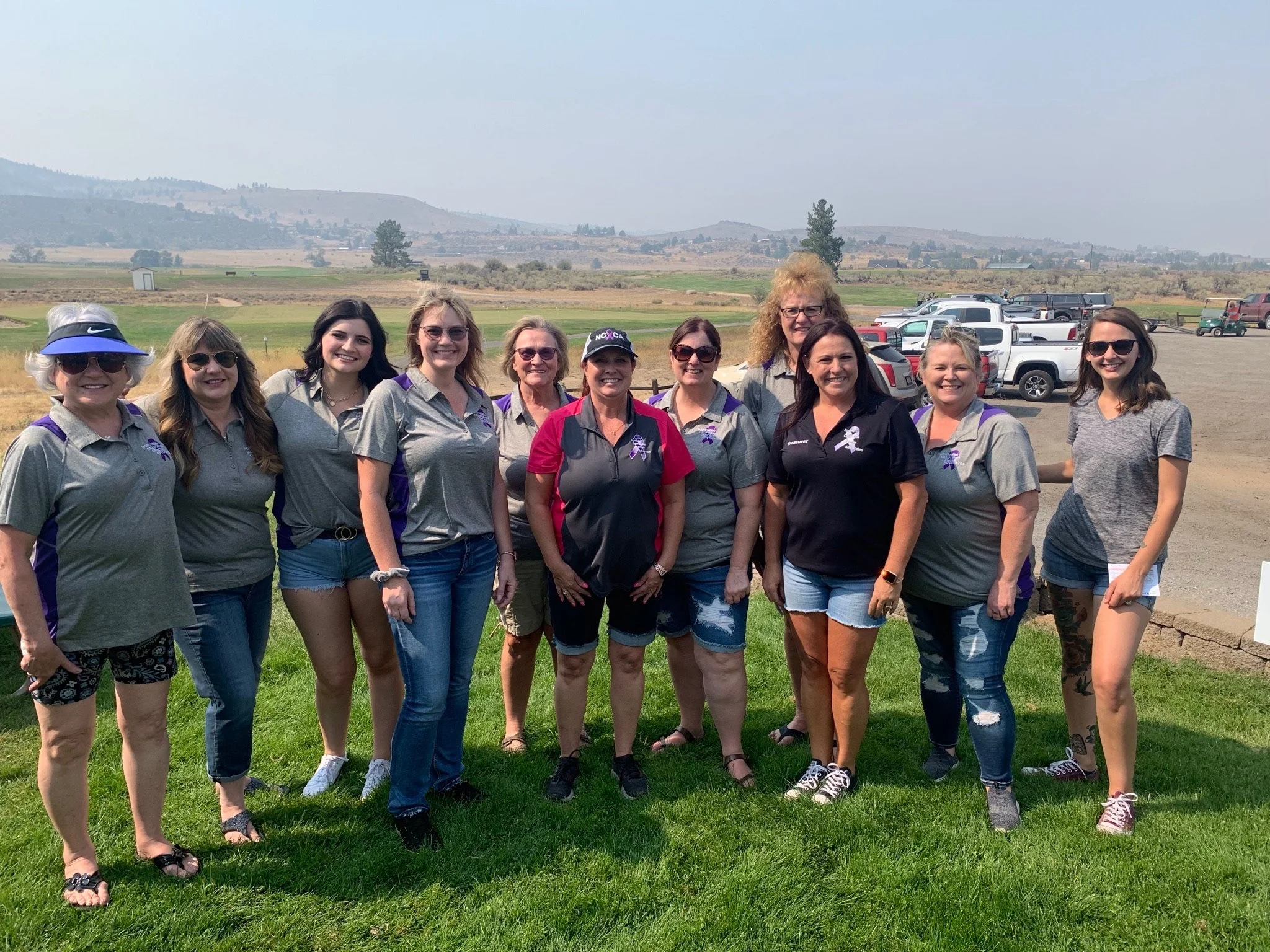 A group of women standing outdoors on grass, smiling, with a rural landscape and parked cars in the background.