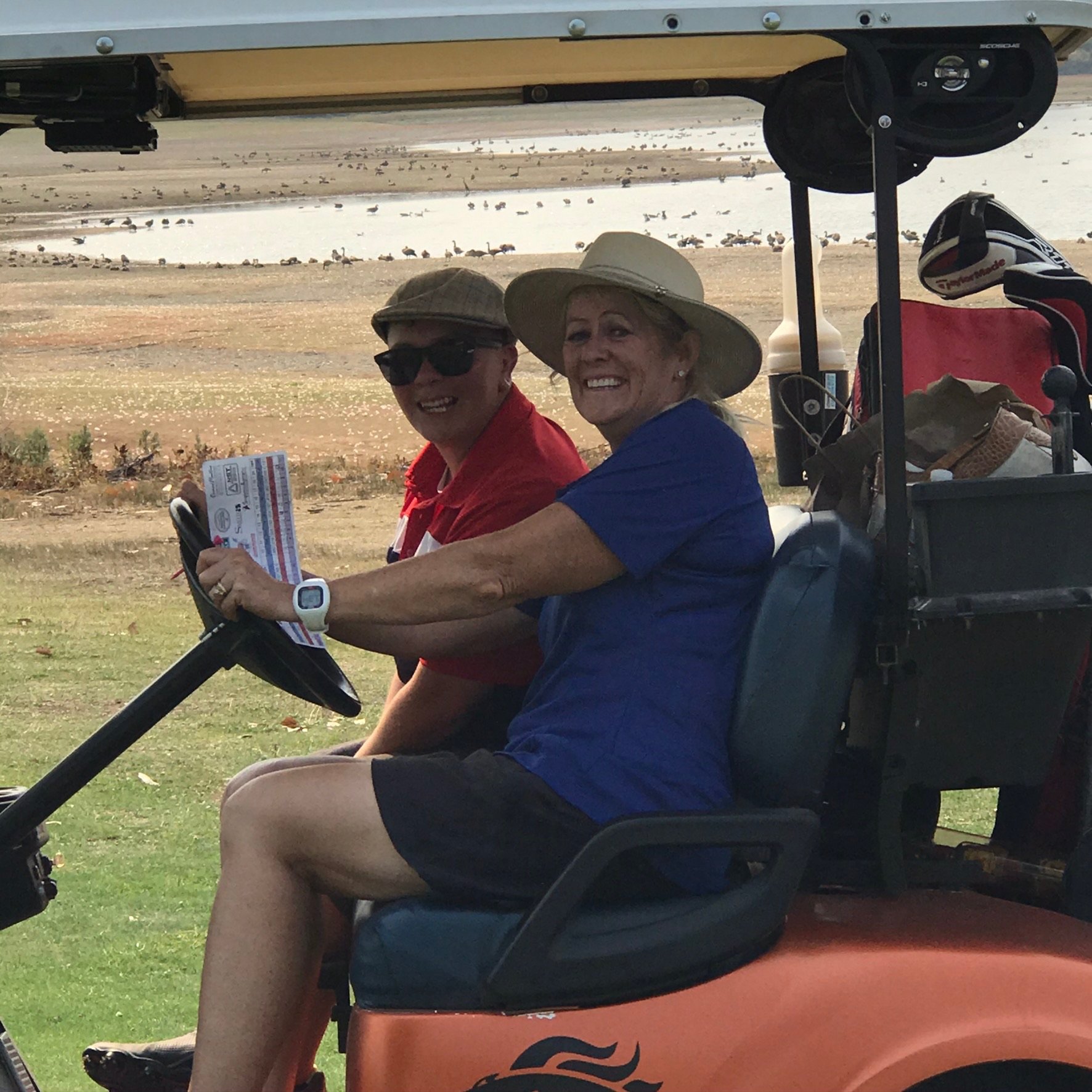 Two smiling women sitting in a golf cart, wearing sun hats, with a golf course and ducks in the background.