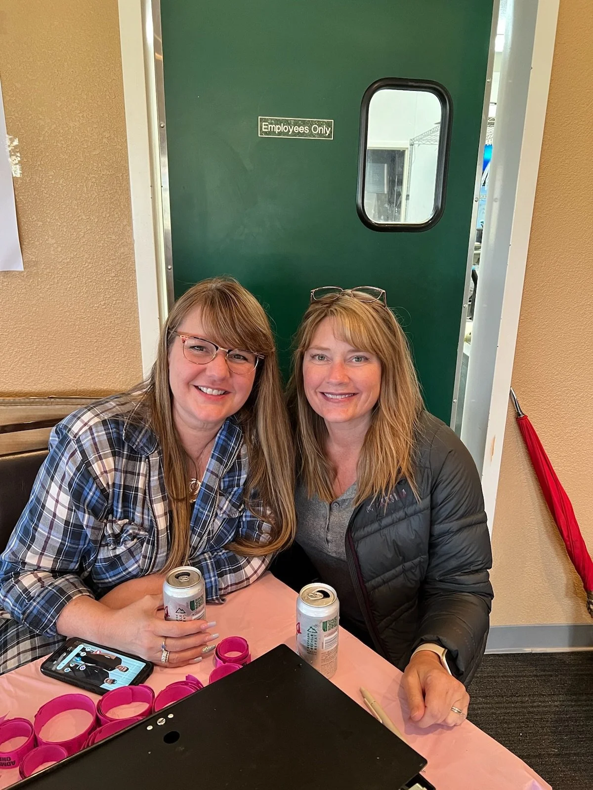 Two women sitting at a table smiling, with drinks in hand and pink wristbands on the table, in front of a green door labeled 'Employees Only'.