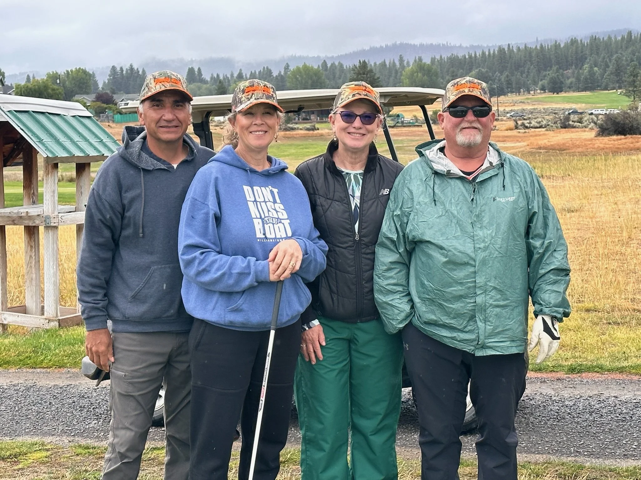 Four people standing outdoors on a cloudy day, wearing camouflage baseball caps and casual clothing, with a golf cart and a rural landscape in the background.