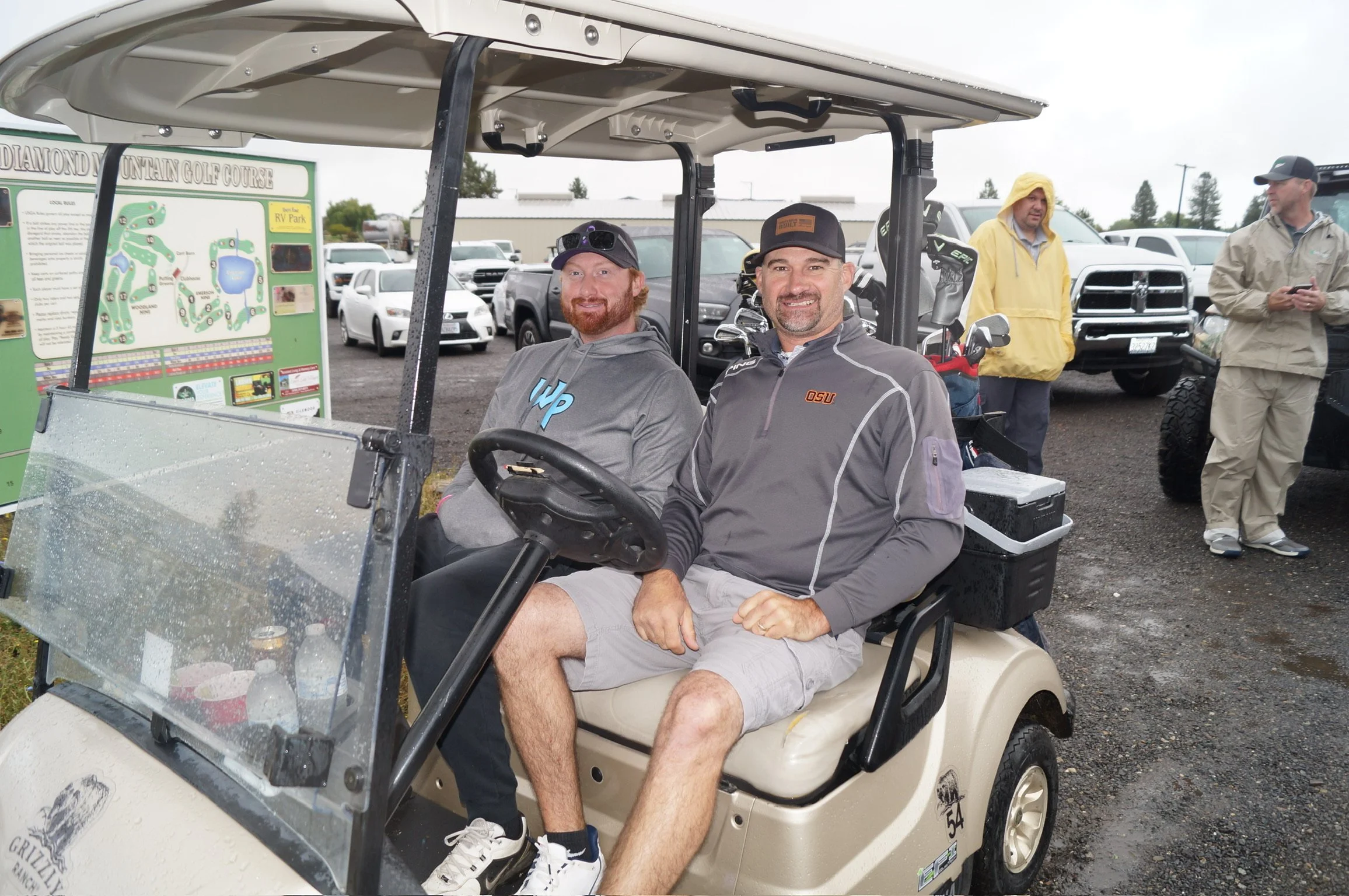 Two men sitting in a golf cart at a golf course, with several other people and vehicles in the background on a rainy day.