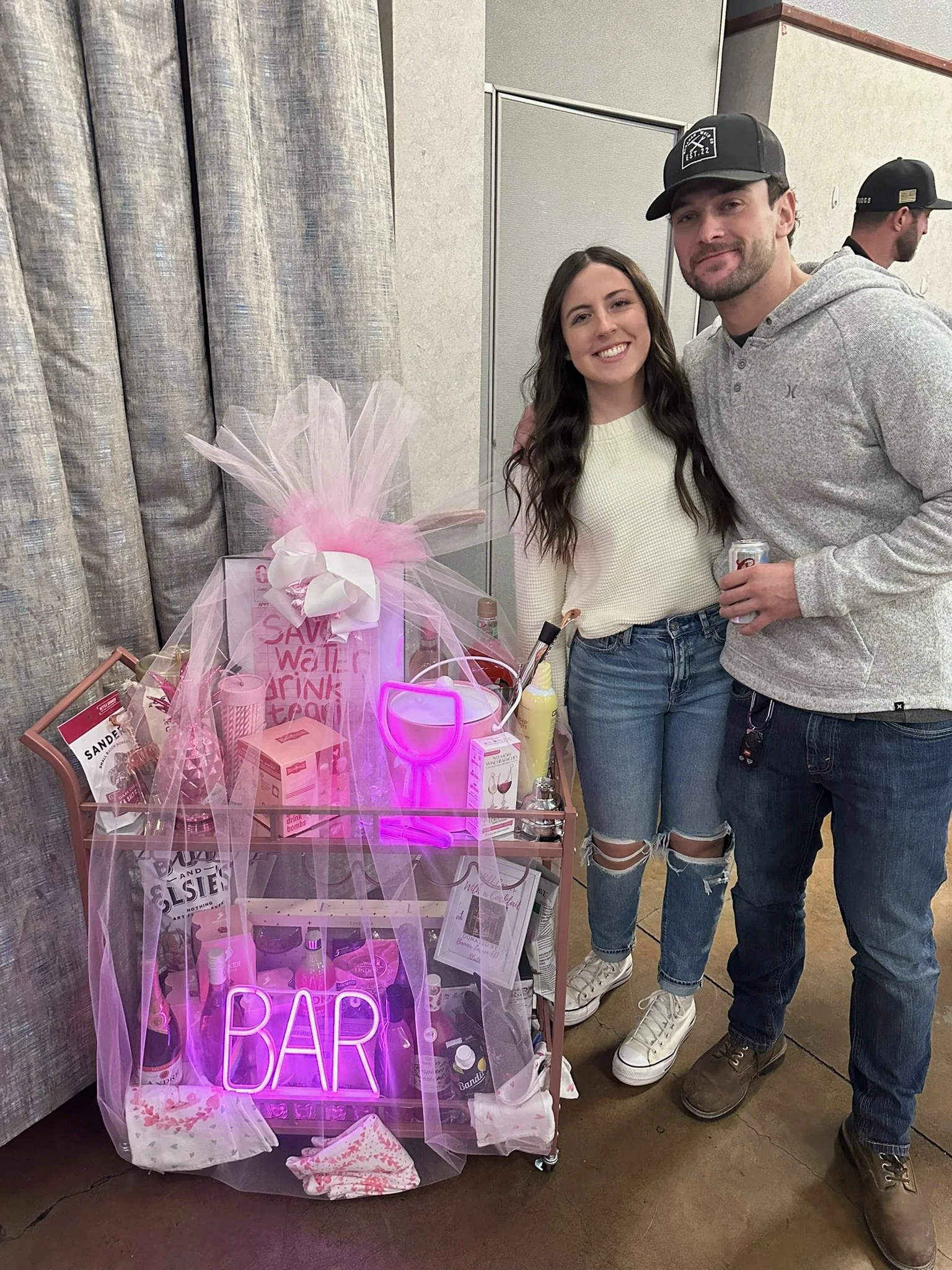 A couple is posing for a photo next to a small pink bar cart decorated with pink tulle, a neon sign that says 'BAR', and various drink items. The woman is smiling, wearing a white sweater and ripped jeans, and the man is smiling, wearing a gray hoodi