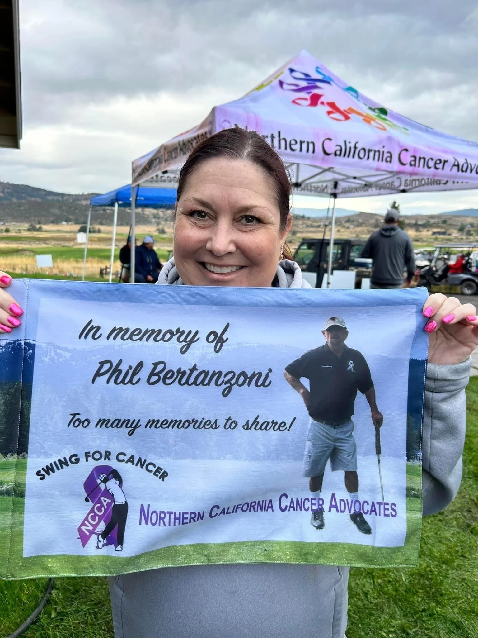 Woman holding a poster in honor of Phil Bertanzoni during a cancer awareness event in a rural outdoor area. The poster features a man with a cane and a message supporting cancer advocates and fundraising.