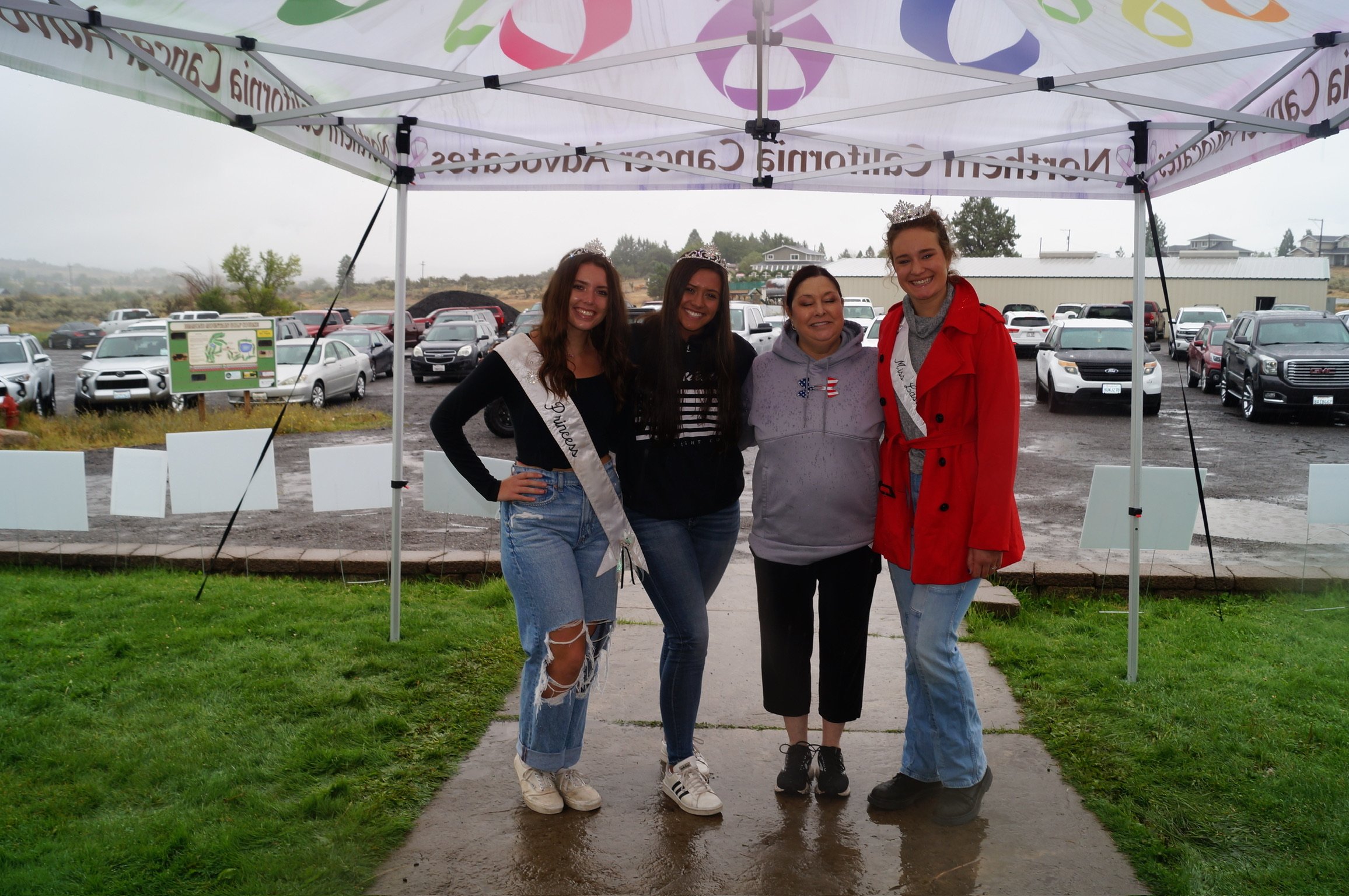 Four women, some wearing tiaras and sashes, stand under a canopy in a parking lot on a rainy day, smiling for a photo.