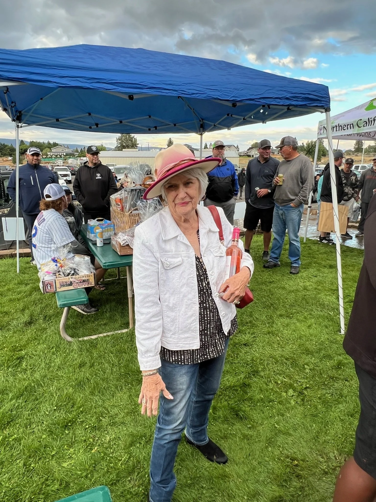 An elderly woman with gray hair and a pink hat stands in front of a group of people under a blue canopy at an outdoor event, holding a bottle and smiling.