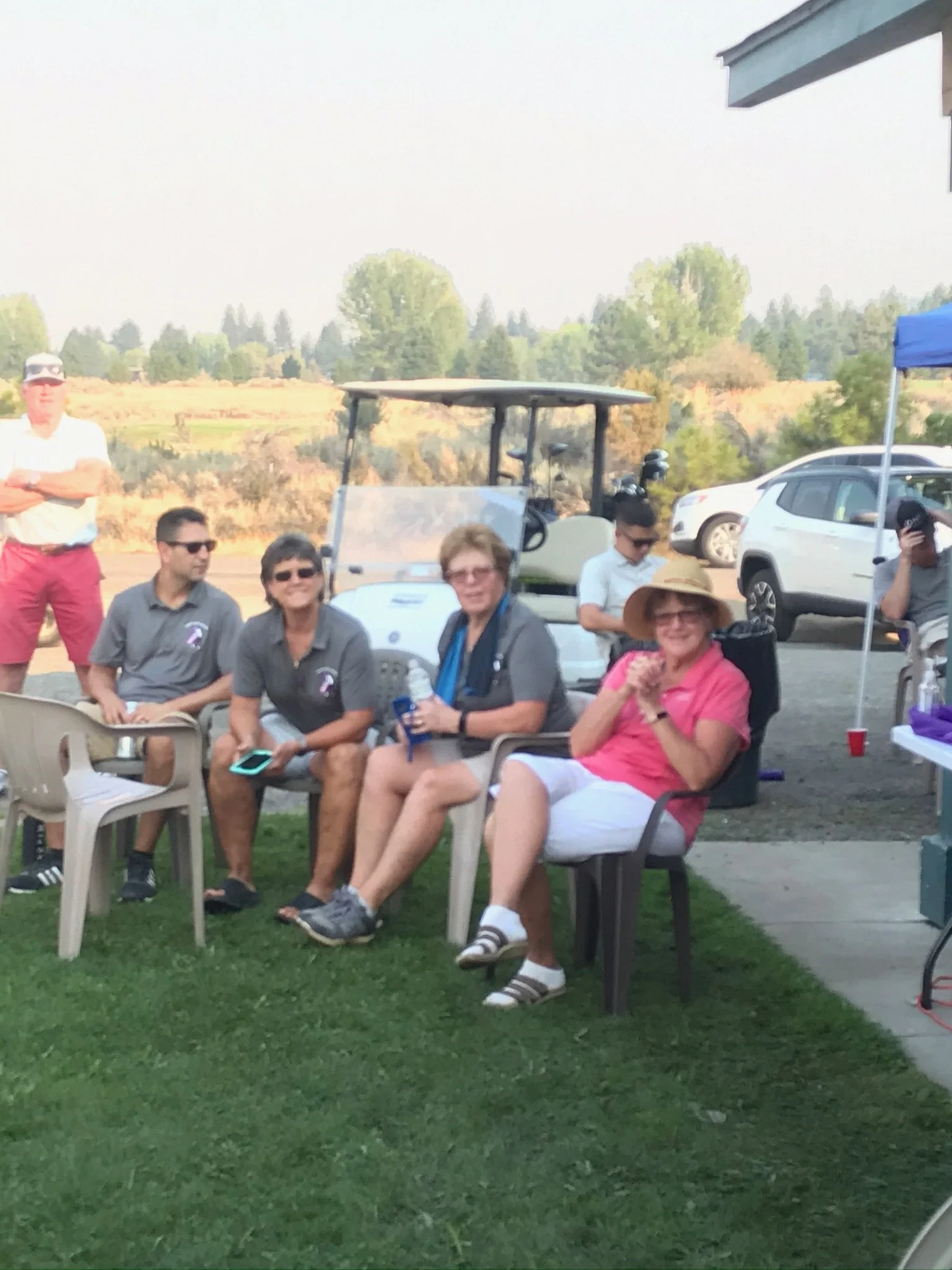 Group of people sitting outdoors on chairs near a golf cart, with a grassy area, trees, and parked cars in the background.