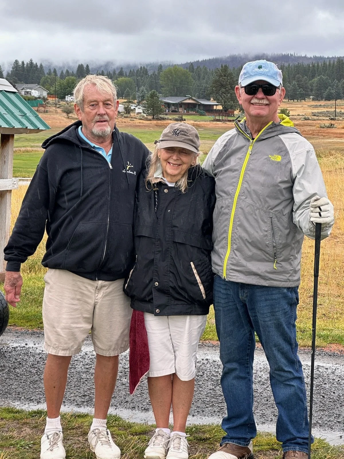 Three older adults standing outdoors on a cloudy day, smiling for the camera. The man on the right is wearing sunglasses, a light gray North Face jacket, and a blue cap, holding a walking stick. The woman in the middle is wearing a black jacket and a