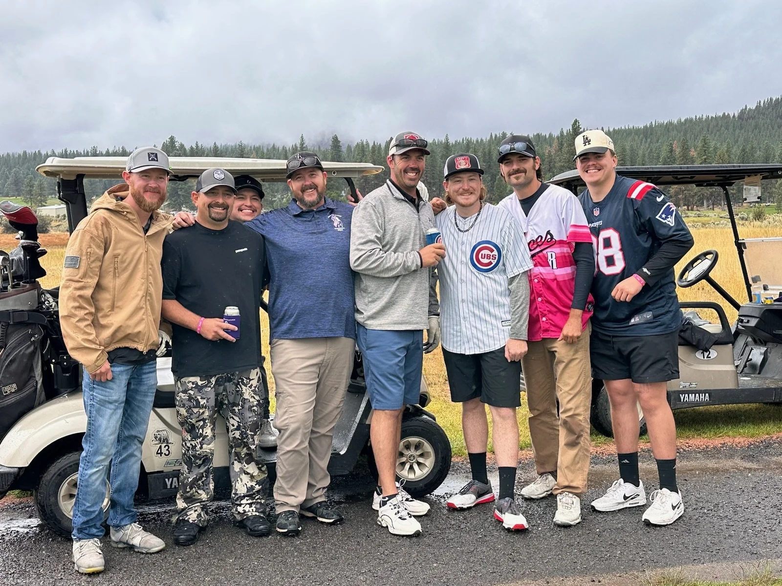 A group of eight men standing together on a golf course, posing for a photo with golf carts behind them and a forested background. They are dressed casually, some in sports jerseys and others in jackets and caps, holding drinks and smiling.