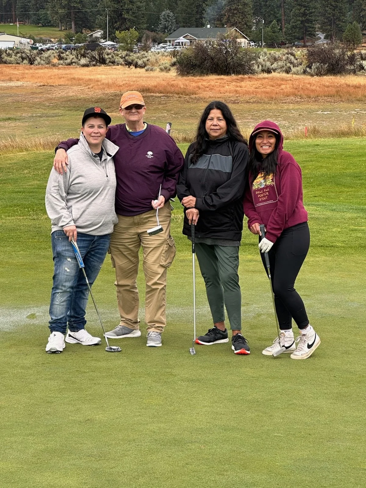Four women standing on a golf course, holding golf clubs, smiling, with a grassy field and trees in the background.
