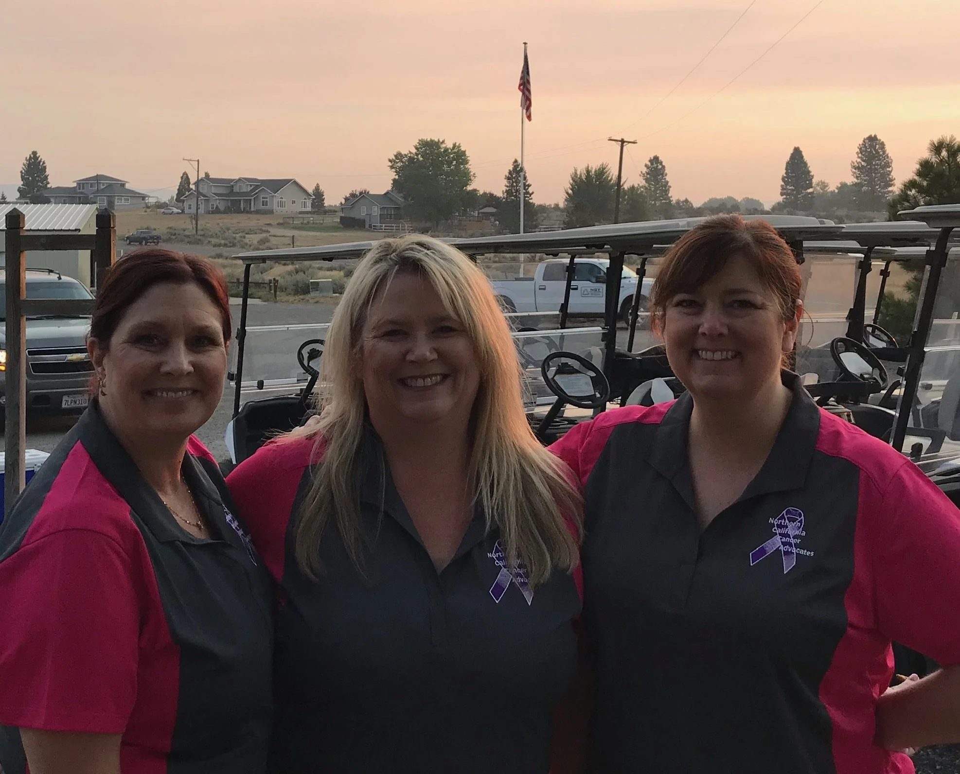 Three women smiling at a gathering, wearing black and pink uniforms with purple ribbons, standing outdoors with golf carts and houses in the background, during sunset.