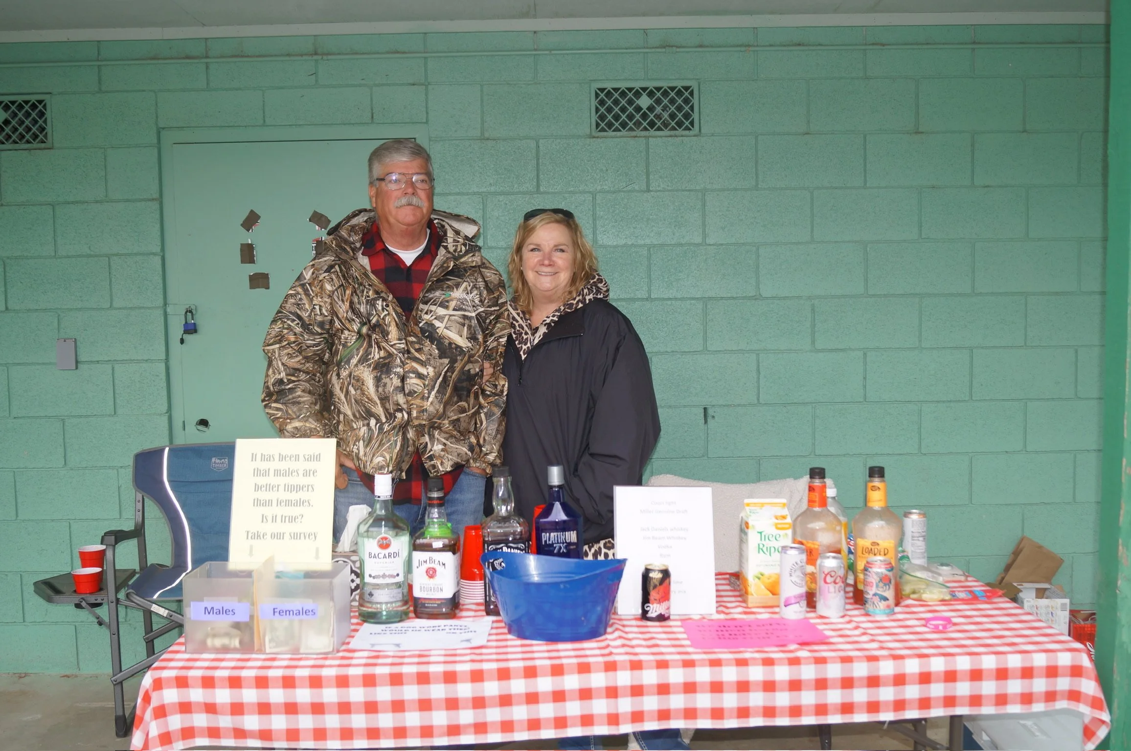 Two people standing behind a table with various drinks and signs, set against a green cinder block wall.