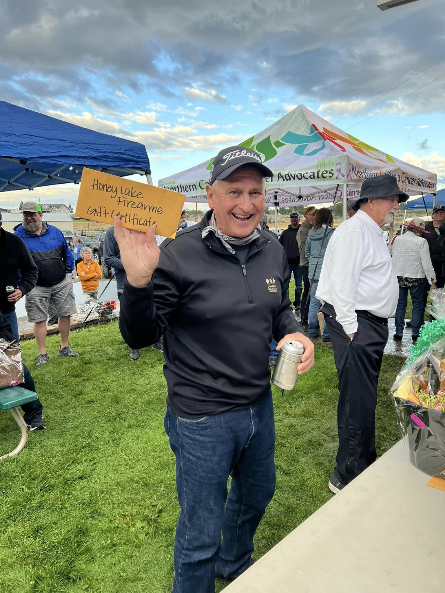 A man smiling and holding a gift certificate that reads 'Honey Lake Firearms Gift Certificate' at an outdoor event with tents and people in the background.