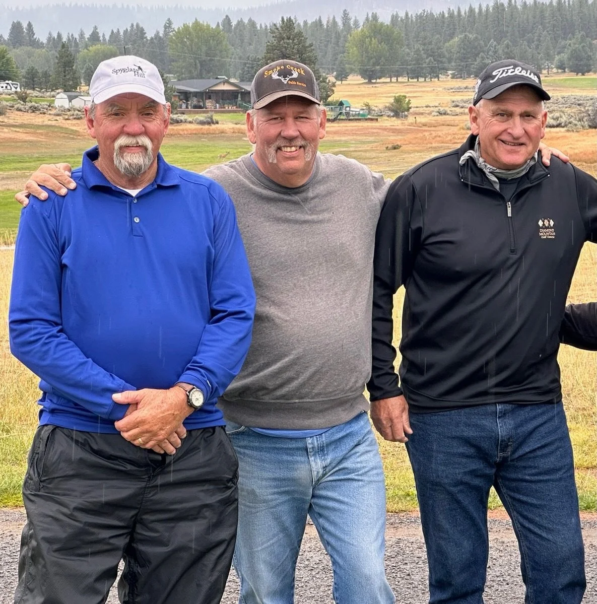 Three men standing outdoors on a cloudy day, smiling, with a rural landscape and house in the background, dressed in casual outdoor clothing including hats and jackets.