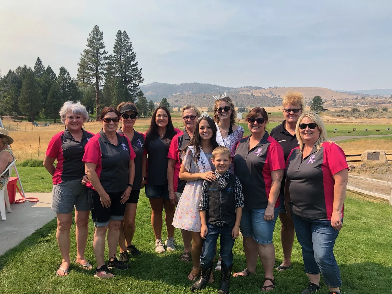 Group of women and children standing outdoors in a grassy area with trees and mountains in the background, celebrating a special occasion.