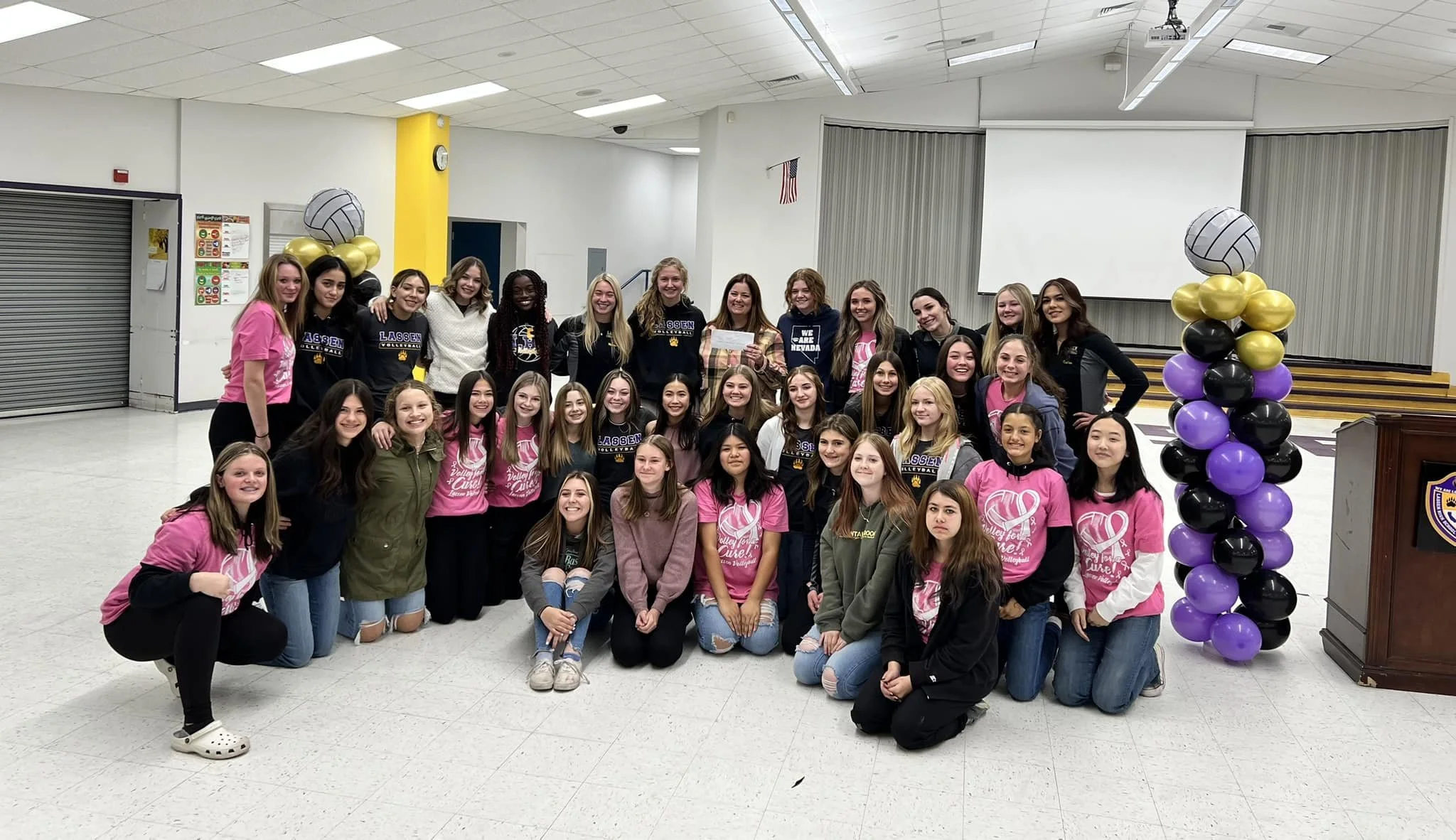 Group of young women gathered indoors for a photo, some wearing volleyball team shirts, with balloon decorations on either side of the group, in a room with white walls and a yellow pillar.