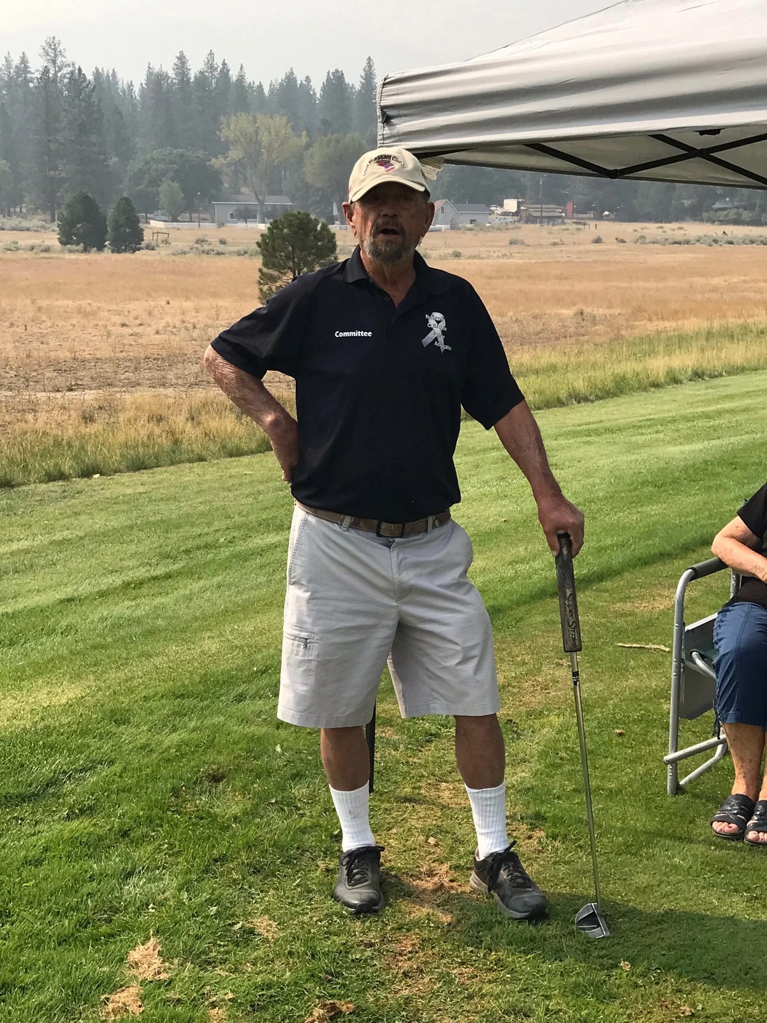 An older man with a beard, wearing a beige cap, black polo shirt with name tag and ribbon, khaki shorts, and black shoes, standing on grass outdoors next to a person in a wheelchair with a golf club.
