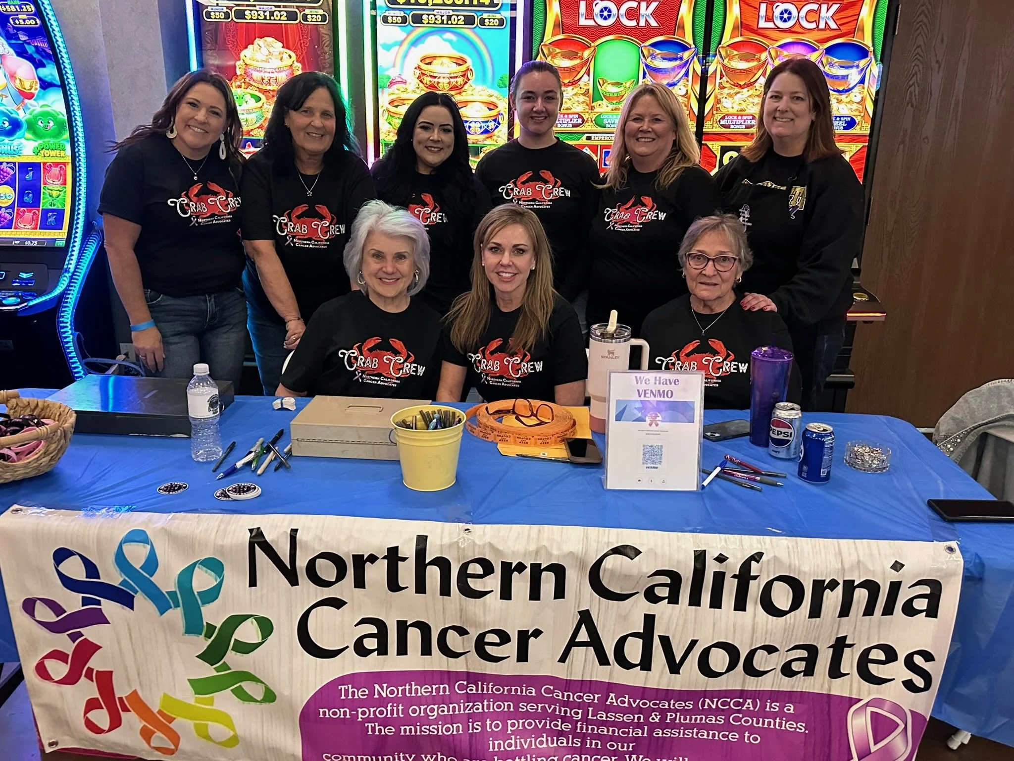 Group of smiling women wearing black 'Crab Crew' t-shirts standing behind a table with promotional materials for Northern California Cancer Advocates. Slot machines are visible in the background.