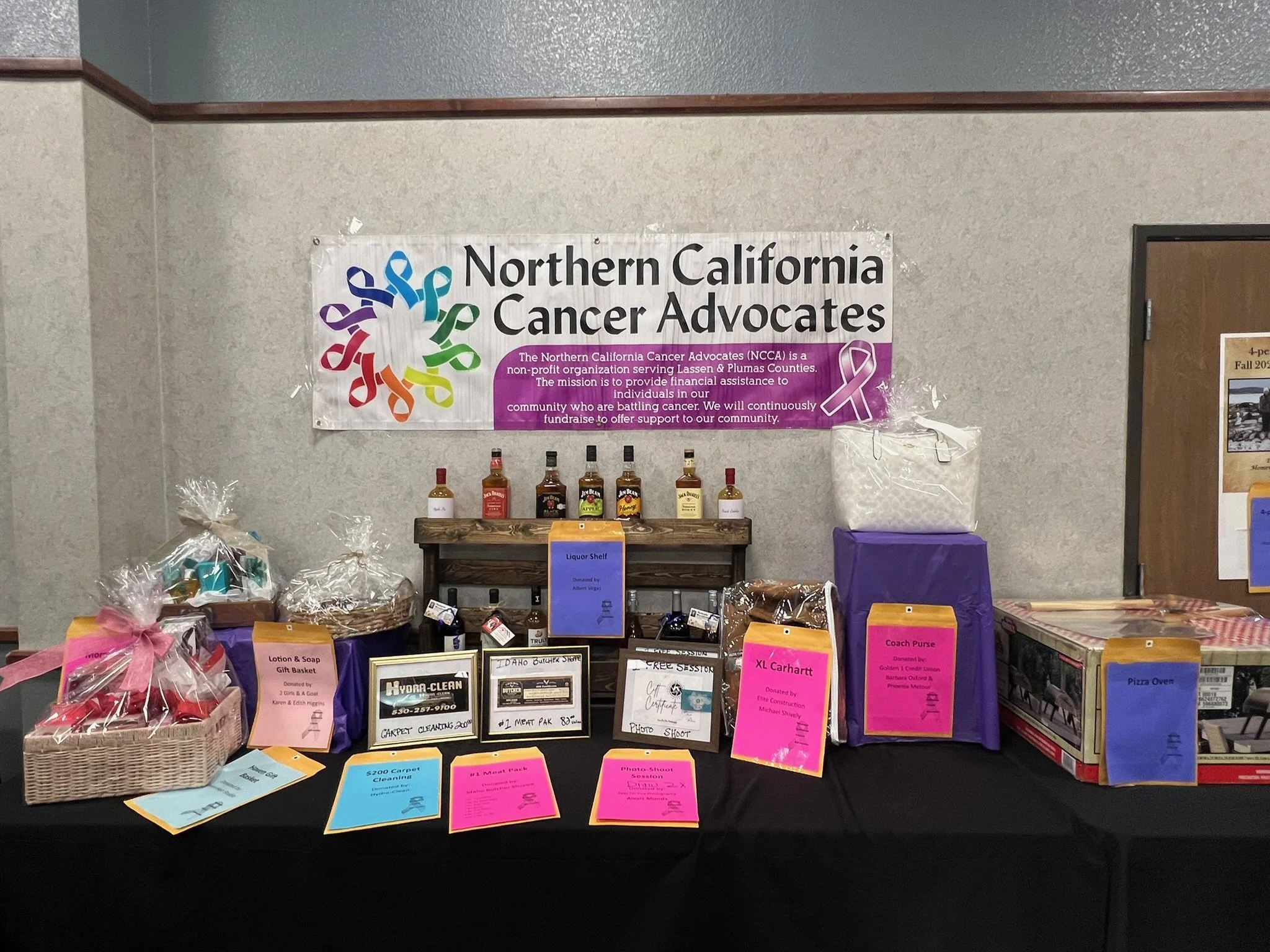 A table at a charity event with various donation and auction items, including bottles of alcohol, gift baskets, and donation notices, with a large Northern California Cancer Advocates banner in the background.