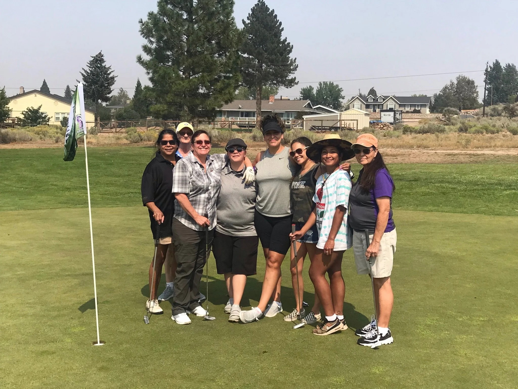 A group of nine women is gathered on a golf course, standing near a flagstick. They are dressed in casual golf attire, wearing sunglasses, hats, and holding golf clubs. The background features trees, residential houses, and a slightly hazy sky.