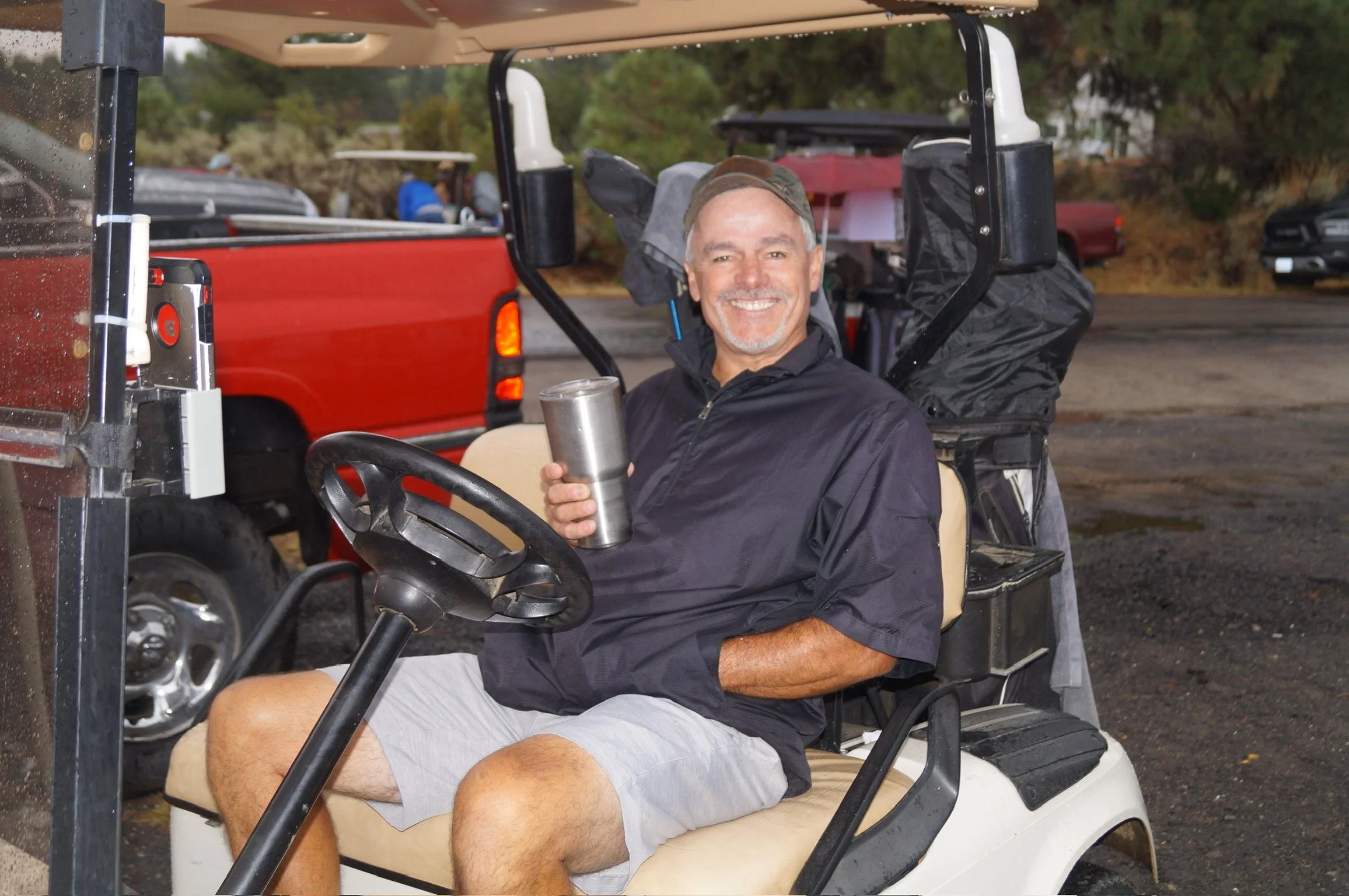 A smiling man sitting in a golf cart, holding a metal tumbler, wearing a cap, black jacket, and shorts, with a golf course in the background.