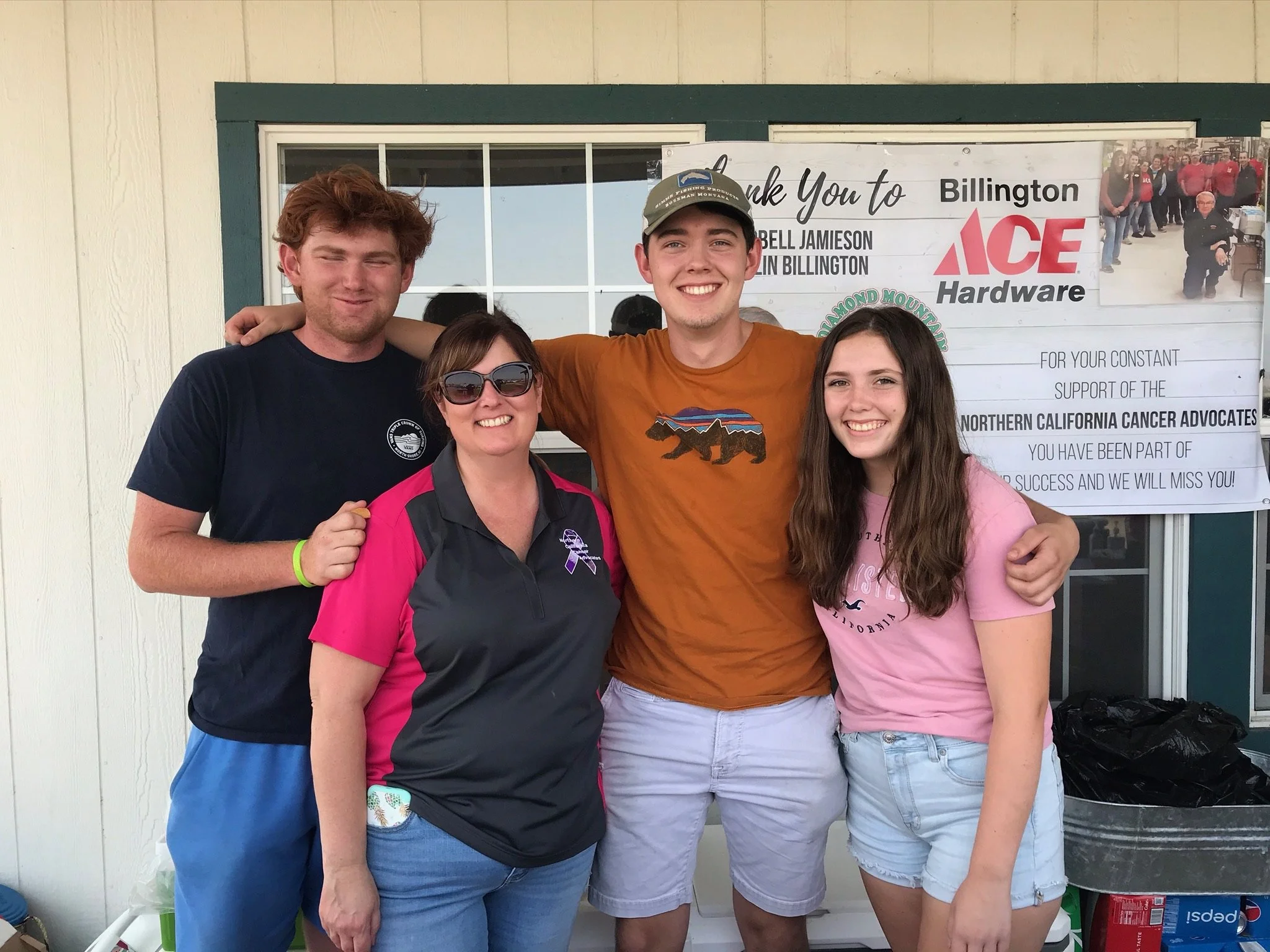Group of four people standing in front of a building and smiling at the camera. Two women and two men, including a young man wearing a baseball cap, a woman in sunglasses, a woman in a pink shirt, and a young woman in a pink shirt with a logo.