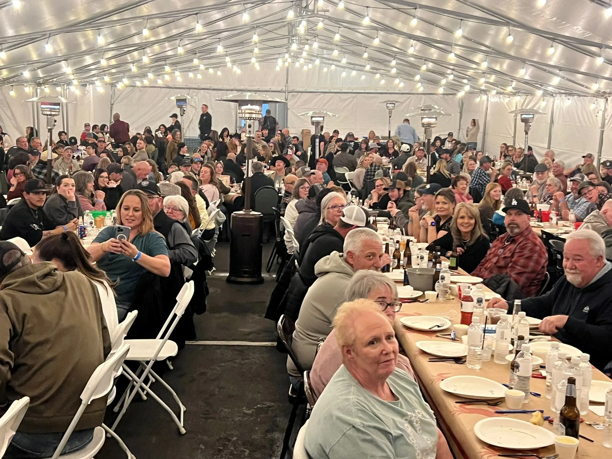 A large gathering of people seated at long tables inside a tent decorated with string lights, enjoying food and drinks.