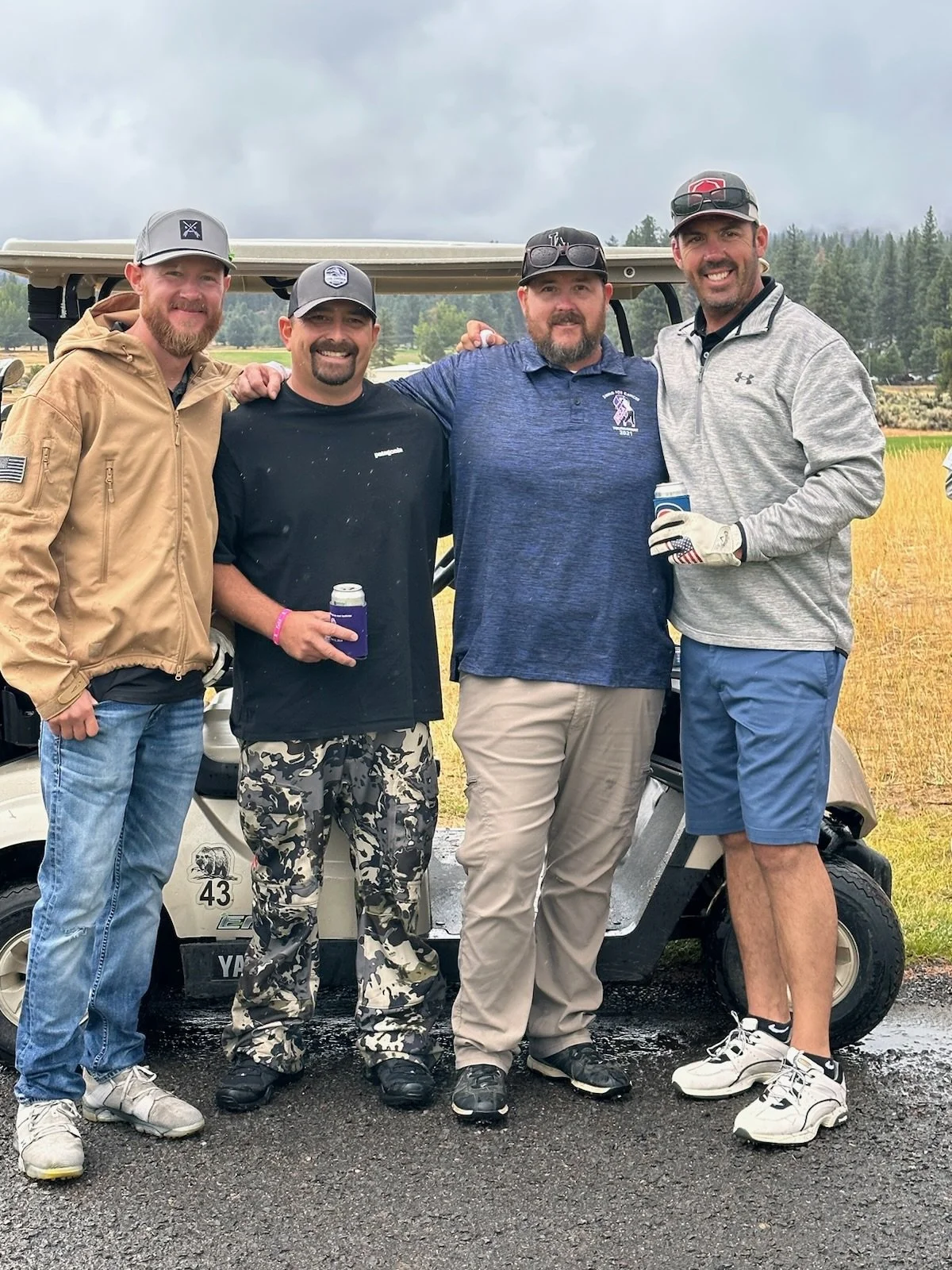 Four men standing together outdoors on a cloudy day, behind a golf cart, with fields and trees in the background. They are smiling and dressed in casual and golf attire.