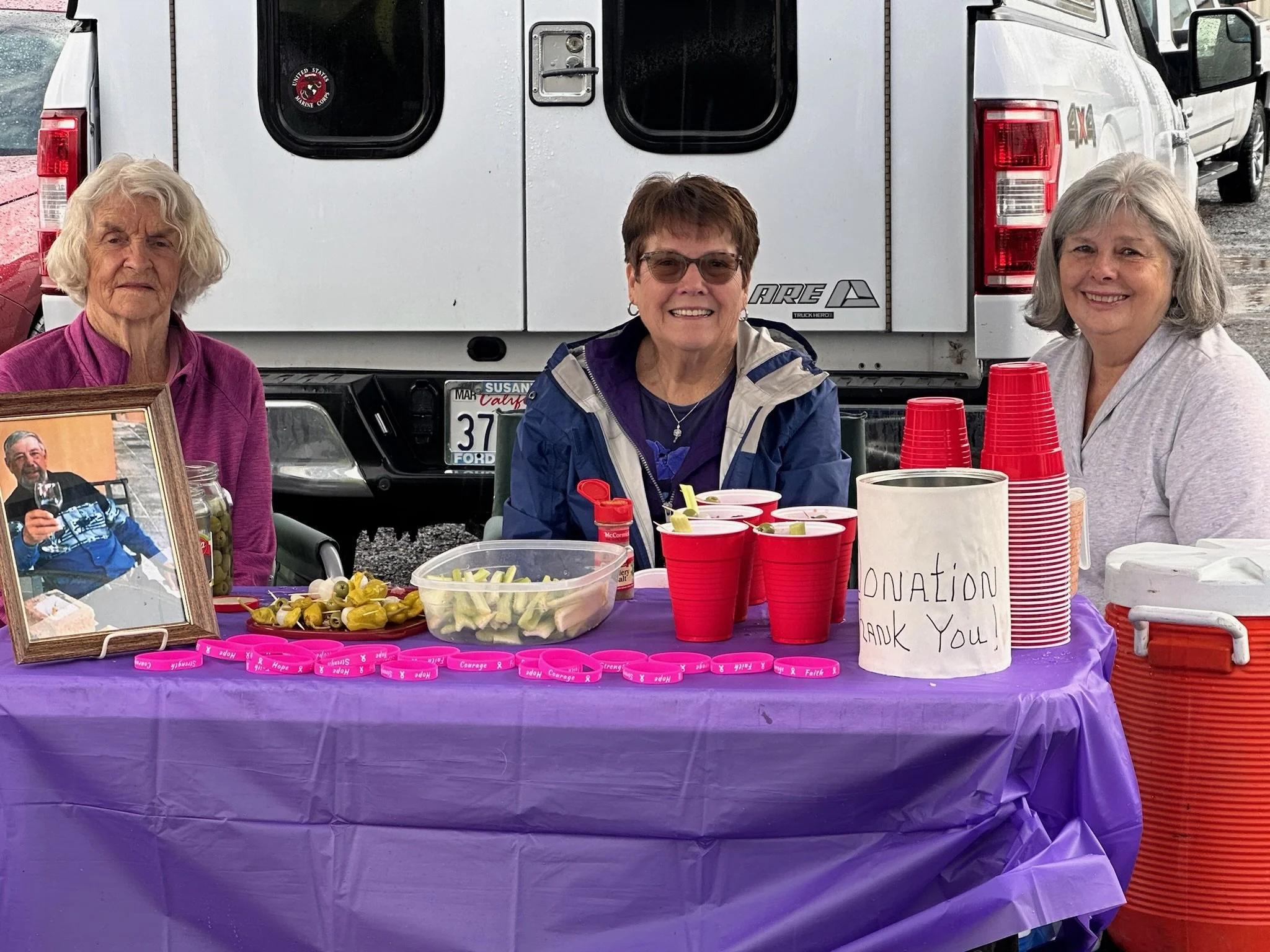 Three women sitting behind a table at a donation drive, with pink bracelets and a sign that says 'Donation'. The table has cups with what appears to be snacks or toppings. There is a framed photograph and a collection container on the table. Behind t
