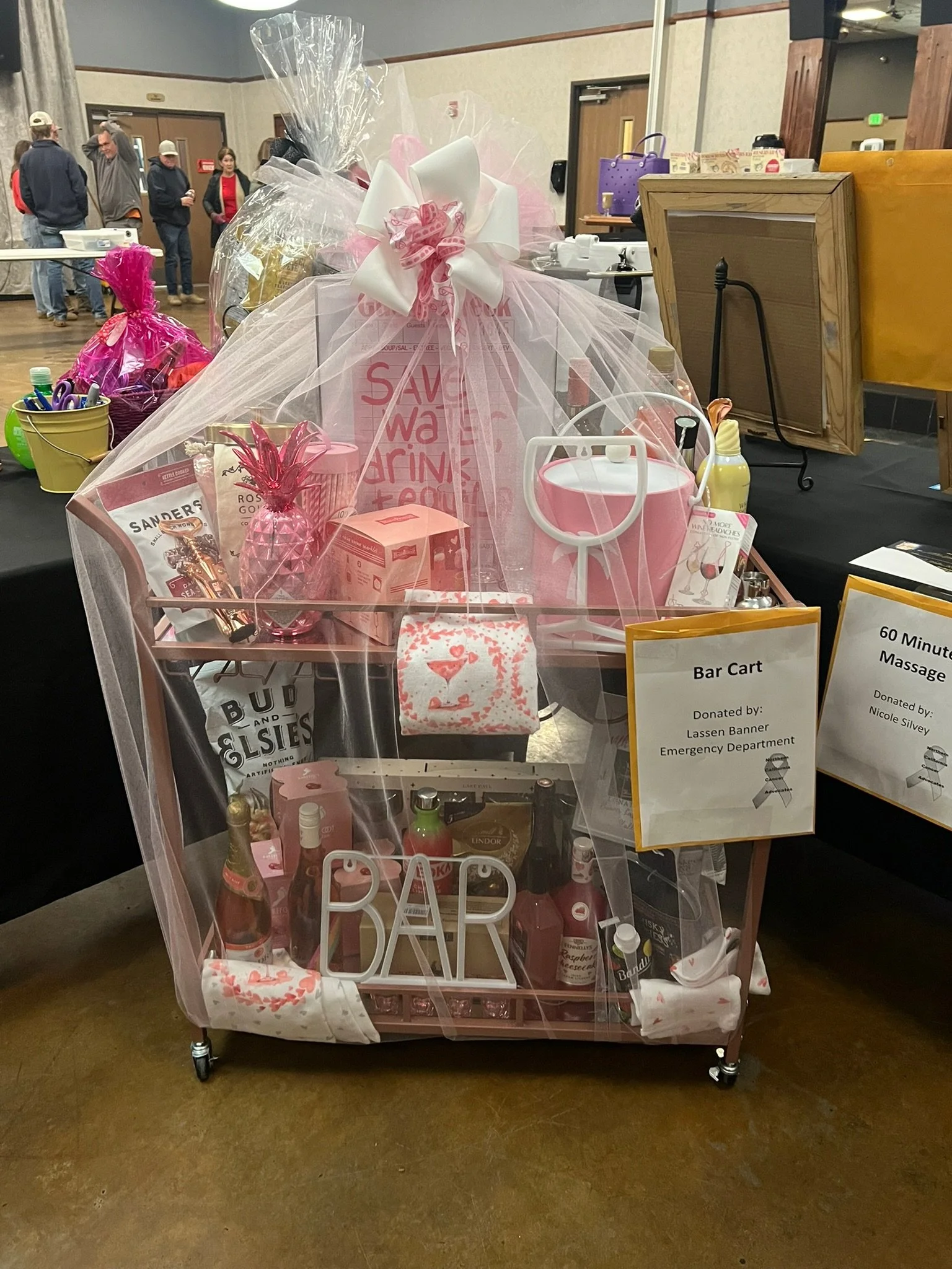 A pink bar cart decorated with pink and white gift wrap, ribbons, and Valentine's Day themed decorations. The cart includes snacks, drinks, and massage gift certificates, and is labeled as donated by Lassen Banner Emergency Department for a charity e