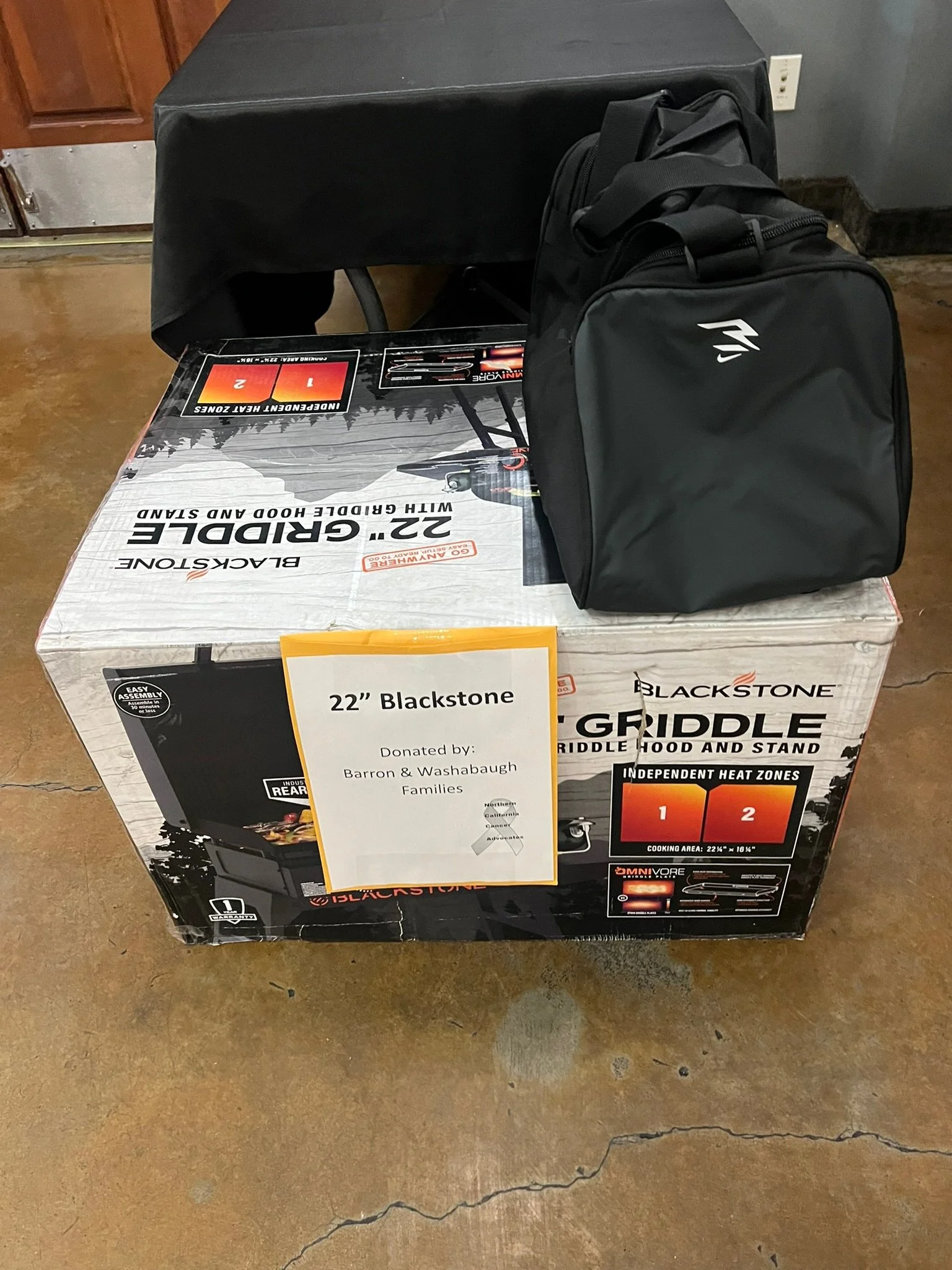 Boxed Blackstone 22-inch griddle with a black bag on top, placed on a concrete floor in a room with wooden cabinets and a black wall.