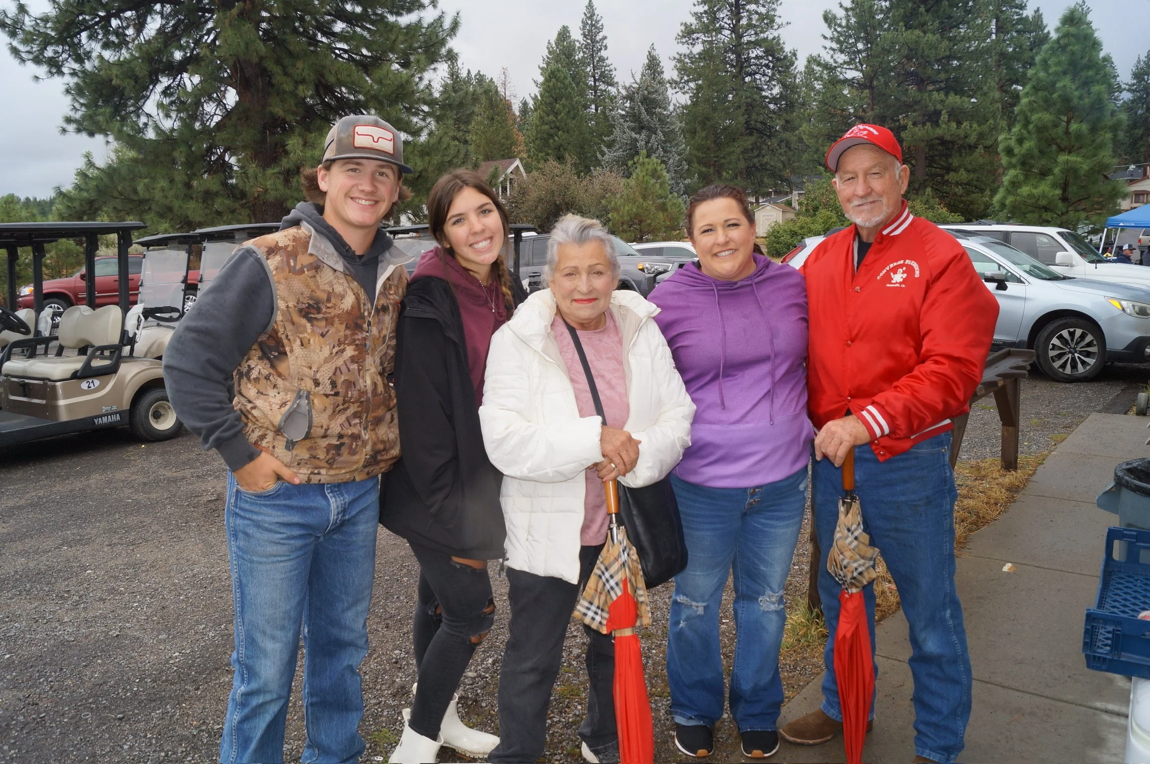 A group of five smiling people standing outdoors in front of a parking lot. They include a young man and woman, an older woman, and a man and woman, all dressed casually. The background has trees and parked cars.