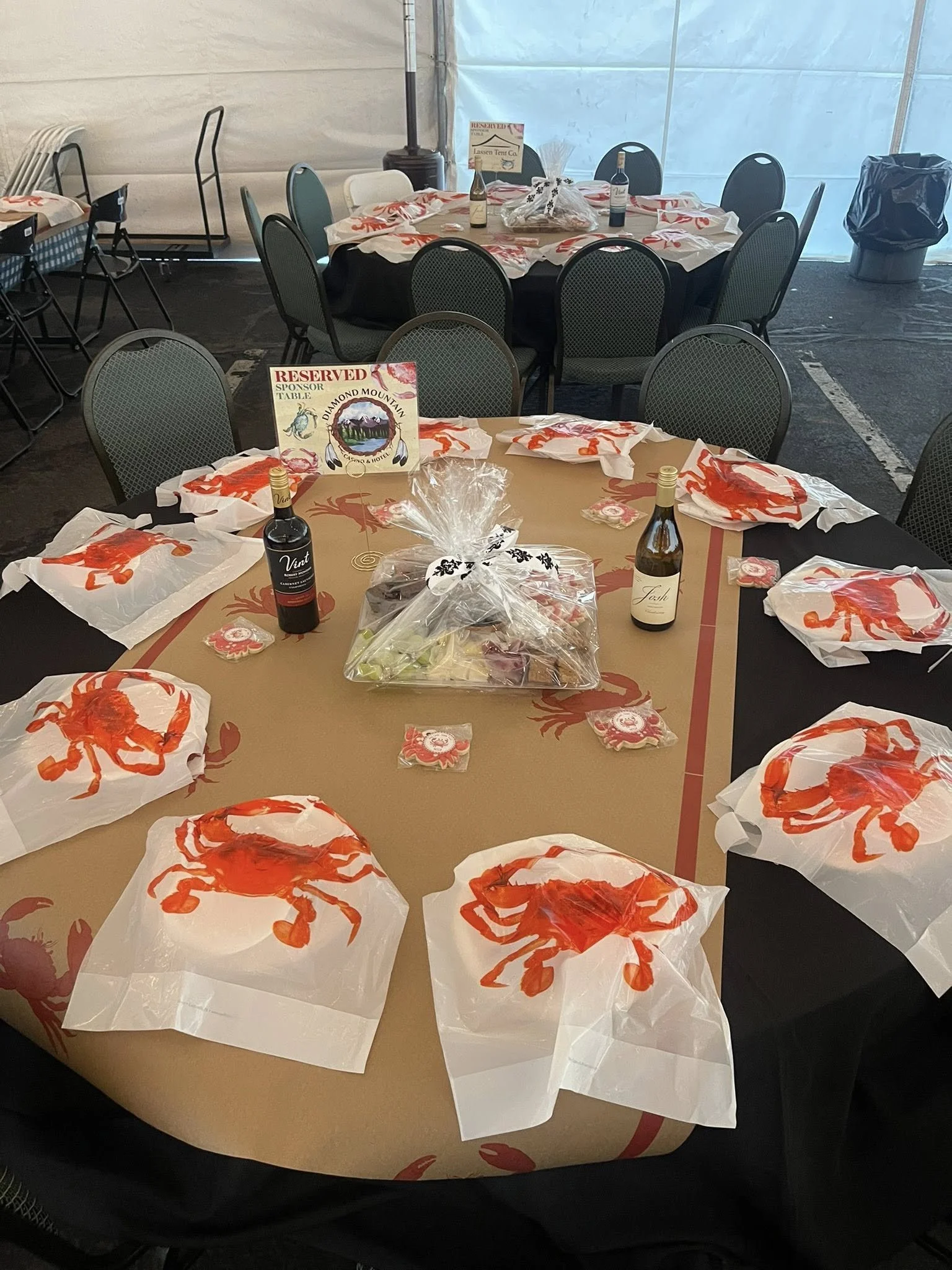 Round tables with black and tan tablecloths decorated with crab and lobster print, set for a seafood event with plates, napkins, and wine bottles; chairs arranged around the tables inside a tent with a door in the background.